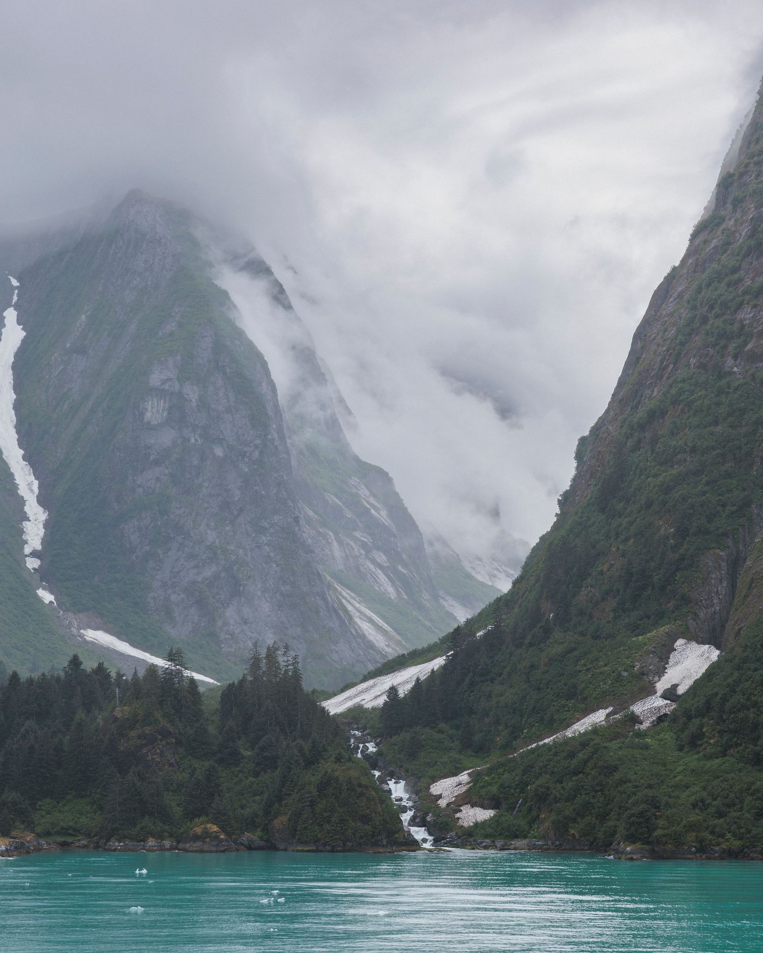 Dramatic snow-capped mountains tower over turquoise glacial waters in an Alaskan fjord.