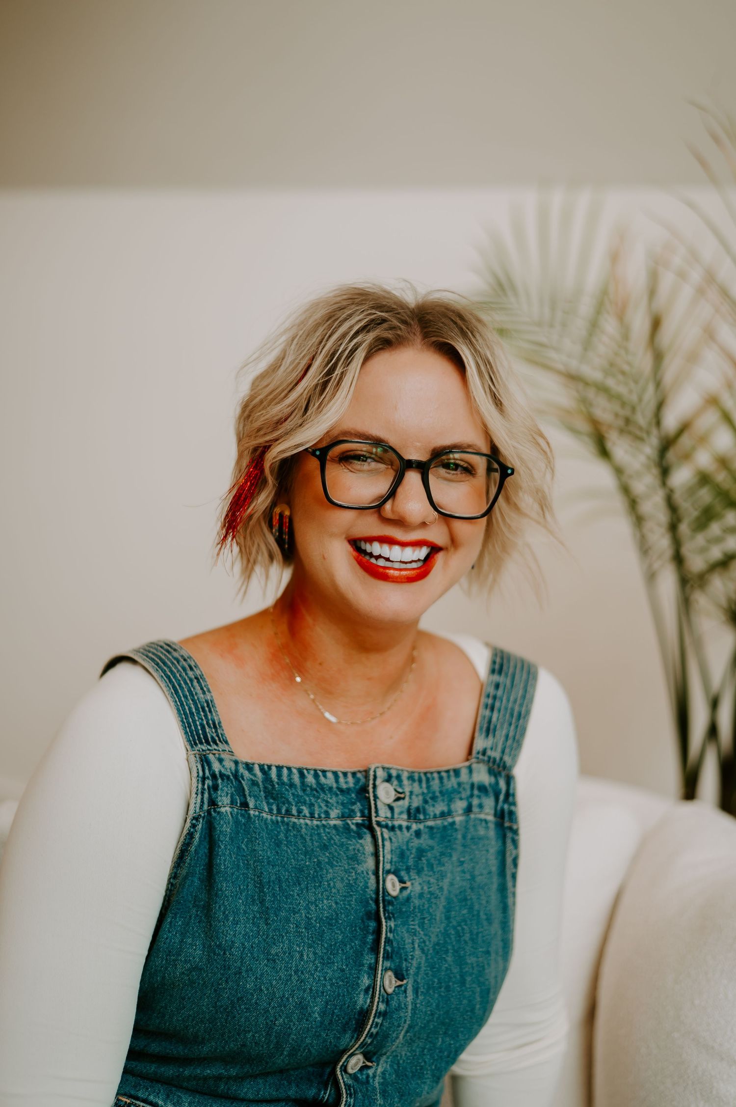 A joyful portrait of someone wearing glasses and denim overalls sitting on a white couch near indoor plants.