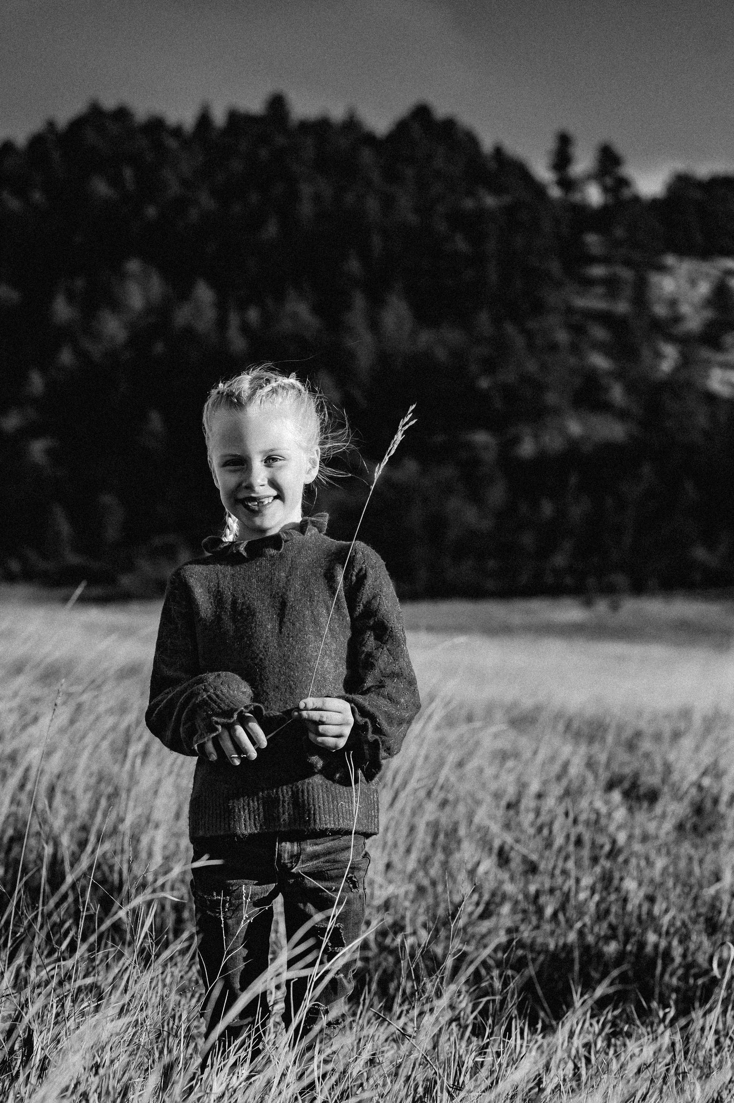 Black and white image of a child sitting in a tall grass field with trees in the background.