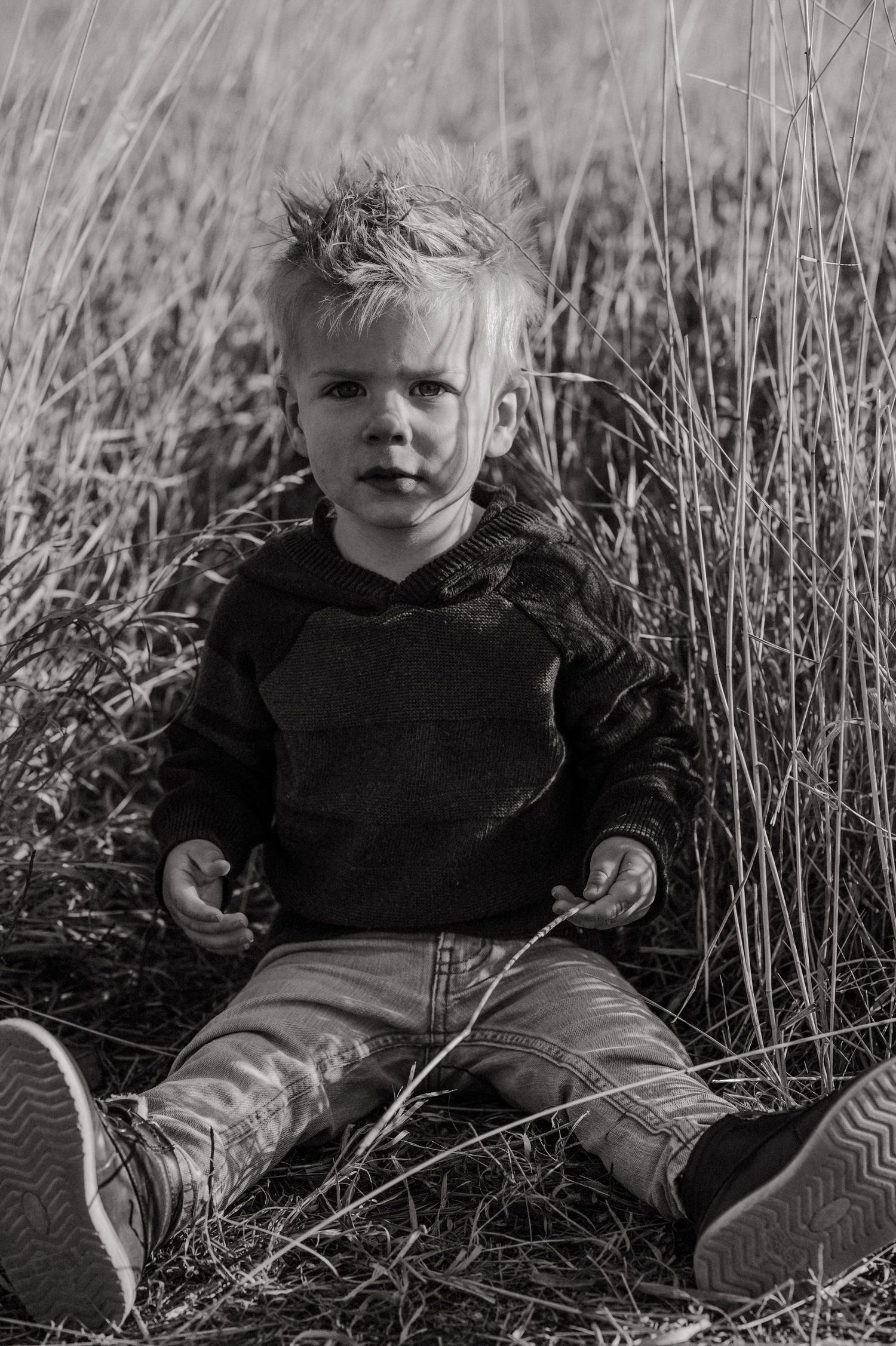 Black and white portrait of a small child sitting in tall grass wearing a dark hoodie and sneakers.