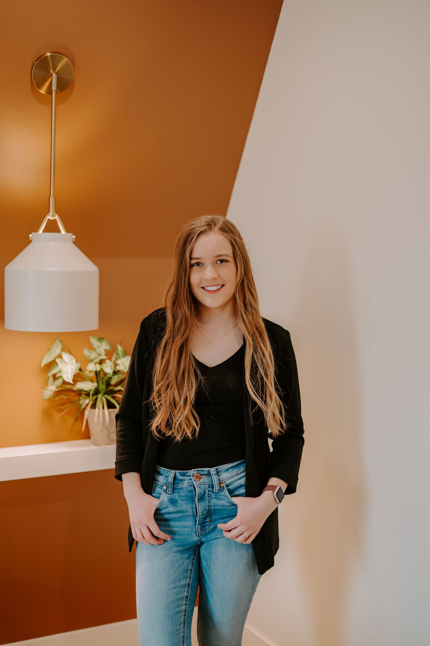 Person wearing black jacket and blue jeans posing against orange accent wall.