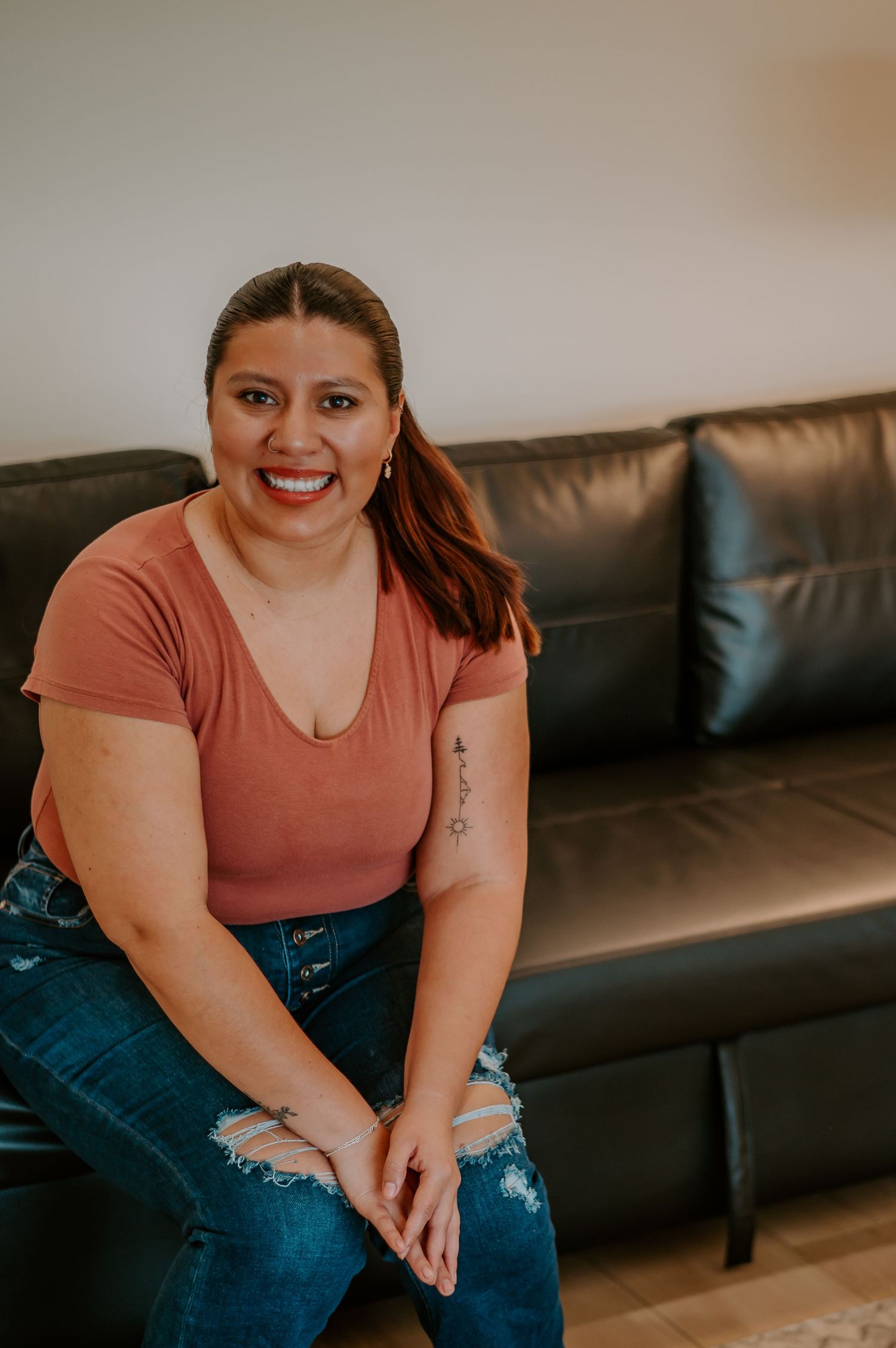 Indoor portrait on a leather couch wearing a coral colored top and distressed jeans.