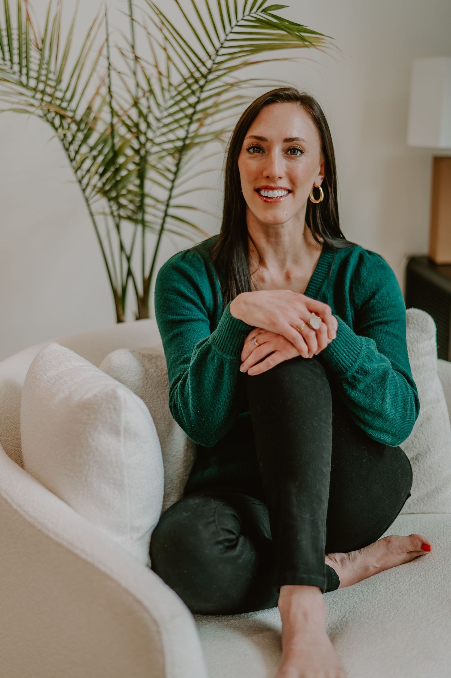 Person in green sweater relaxing on white couch with palm plant in background.