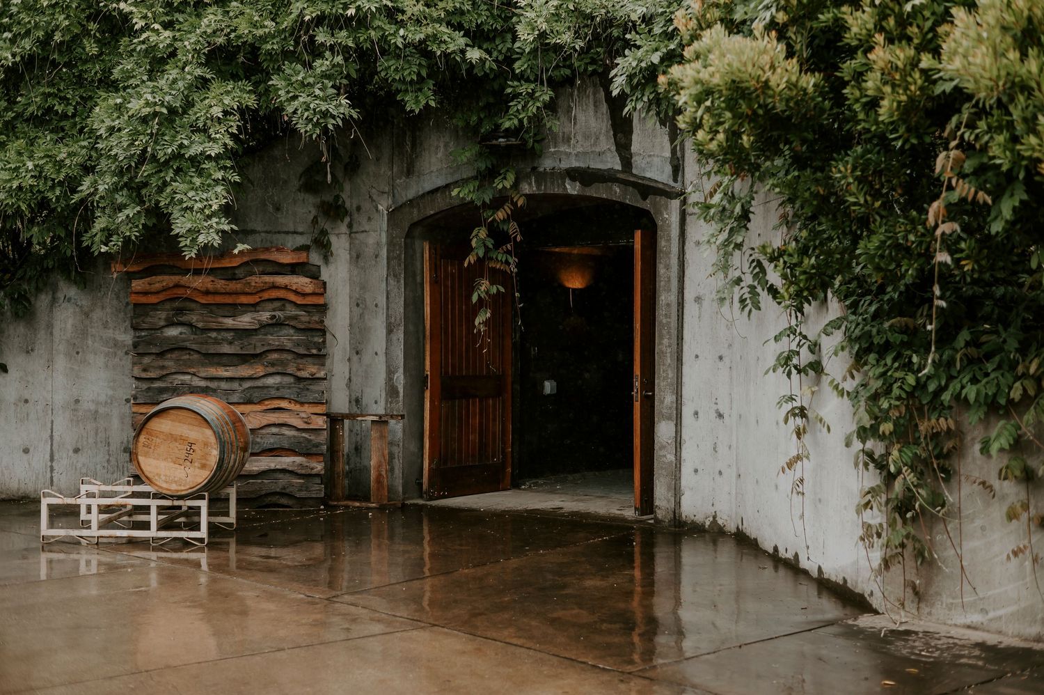 Rustic winery entrance with wooden barrel, stacked wood wall art, and ivy-covered concrete facade with arched wooden door.