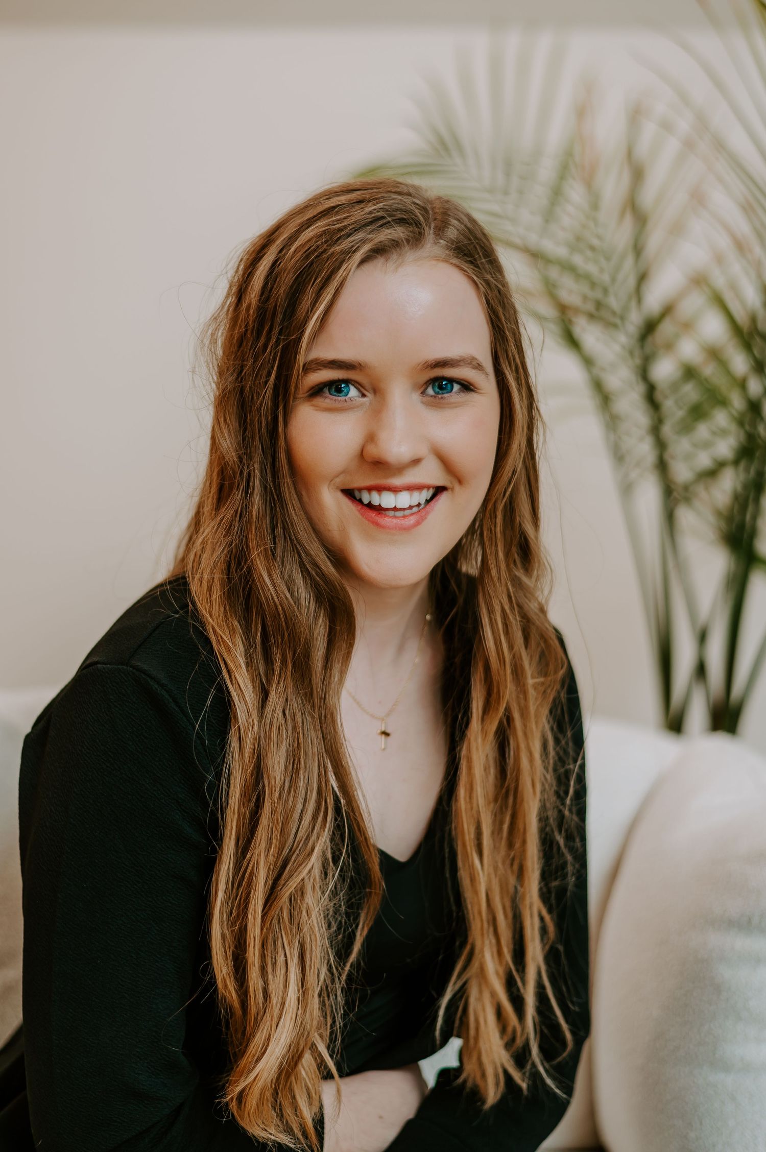 Person in black outfit smiling against white wall with indoor plants in background.