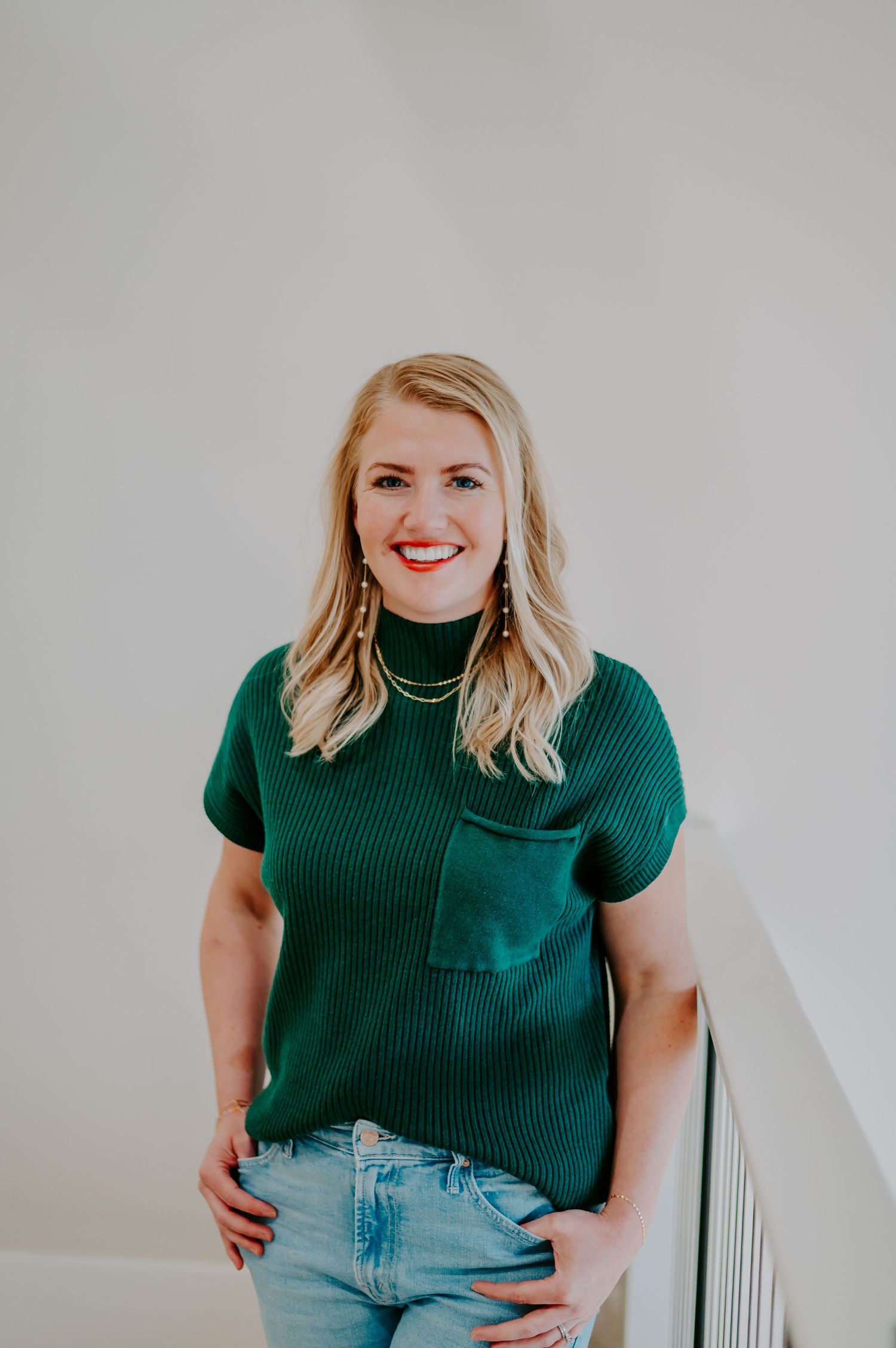 Person wearing green mock neck top and jeans smiling against white background.