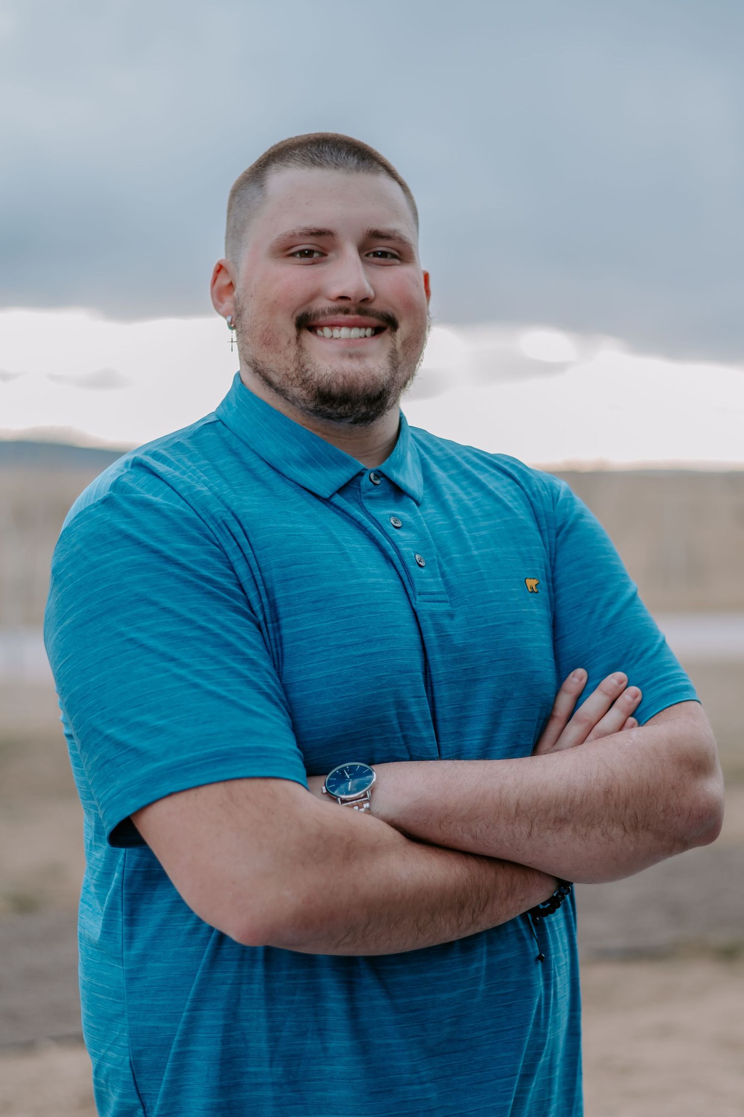 Outdoor portrait in turquoise polo shirt with mountains and cloudy sky in background.
