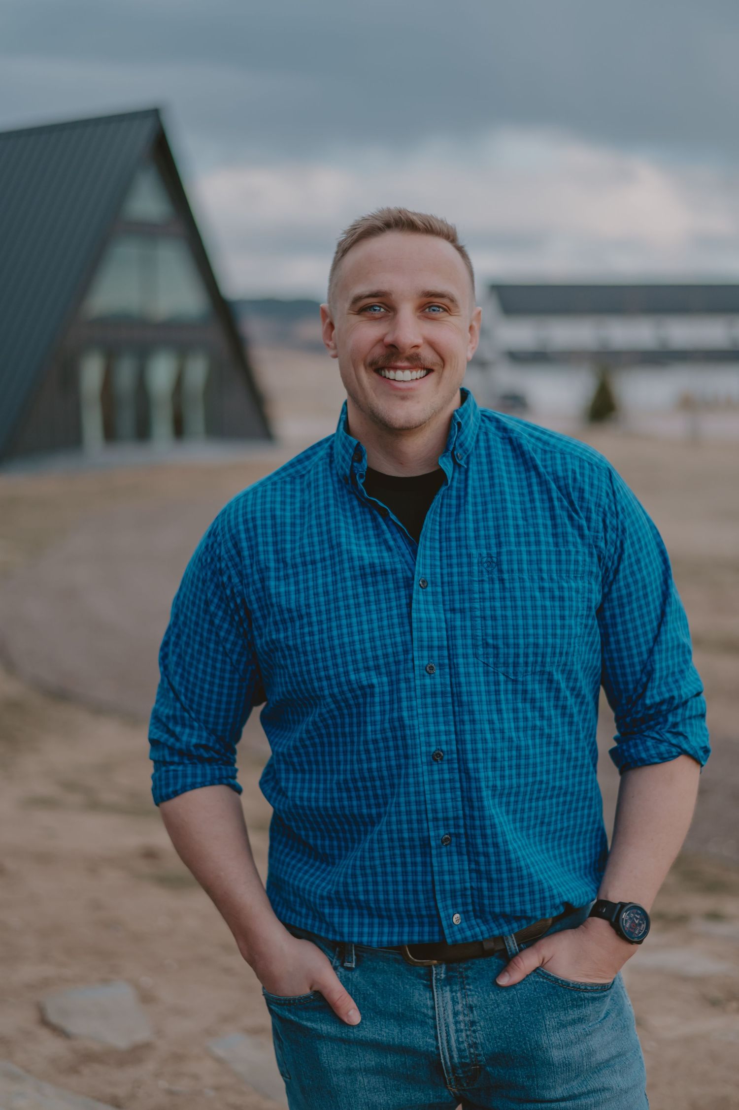 Person in a blue checkered shirt smiling and posing on a ranch with a frame buildings in background.