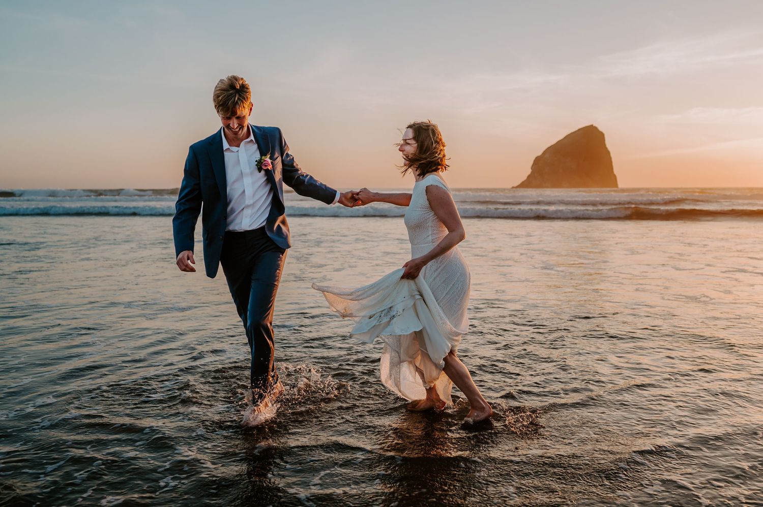 A couple in wedding attire who jumped into the ocean and are laughing together at sunset on the beach in Oregon