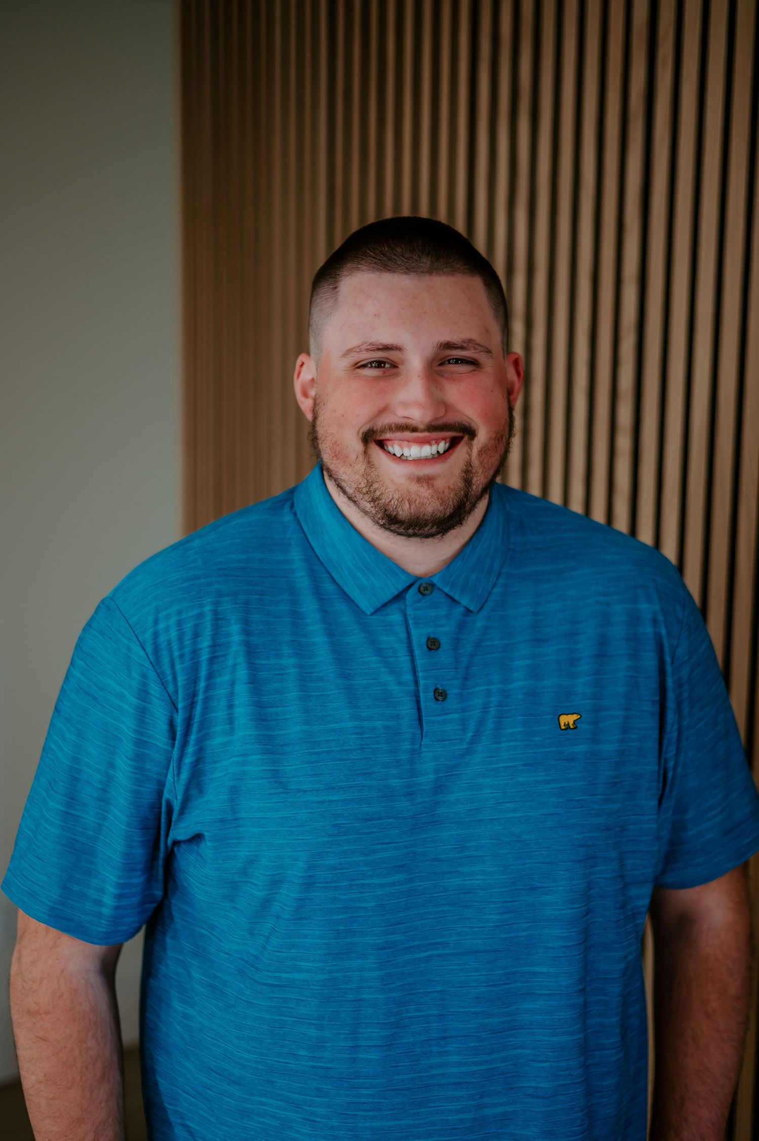 Indoor portrait of someone in a textured blue polo shirt against wooden slat wall background.