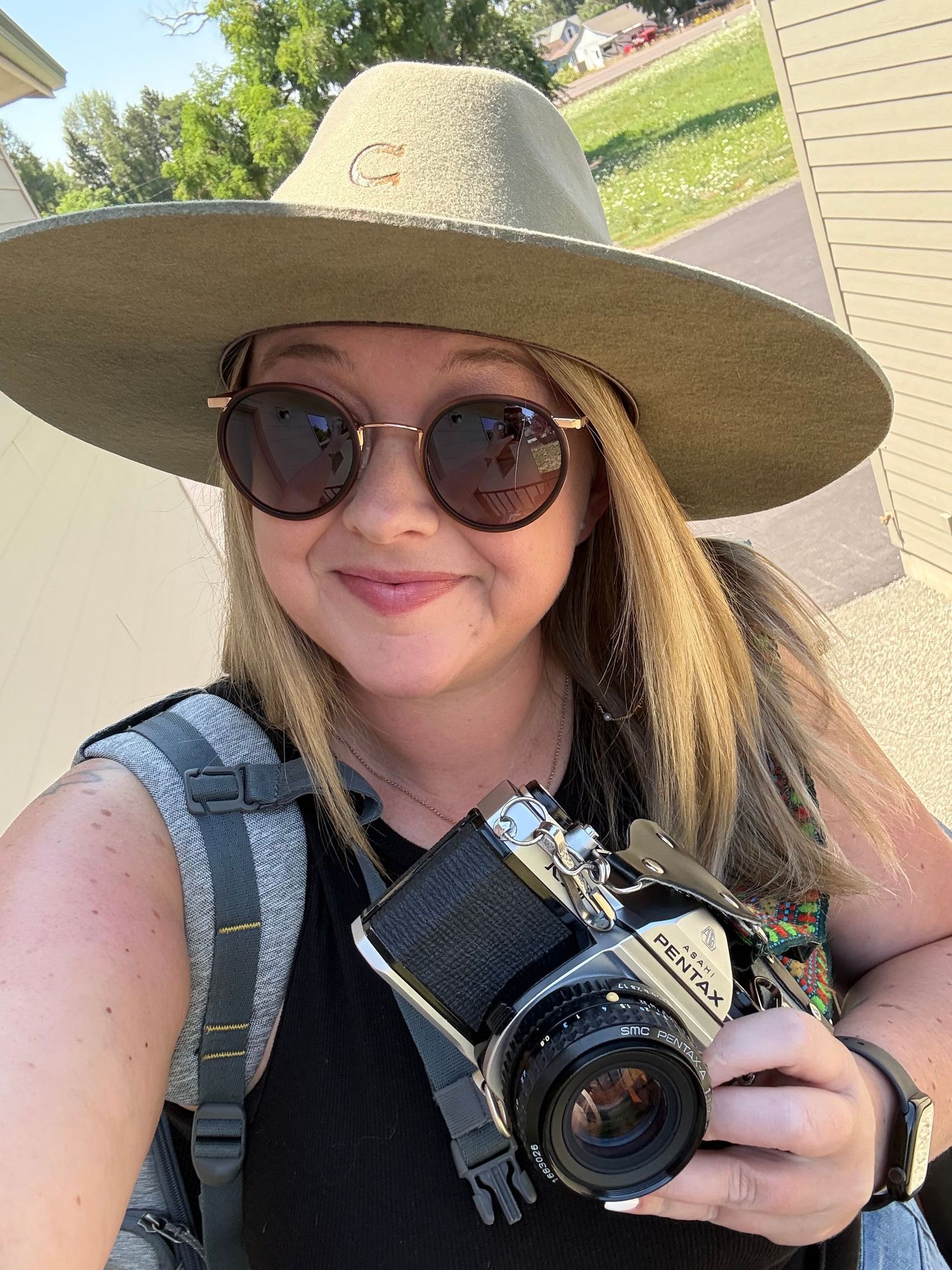 Woman in wide-brim hat and sunglasses holding a vintage Pentax film camera, taking a selfie outdoors.