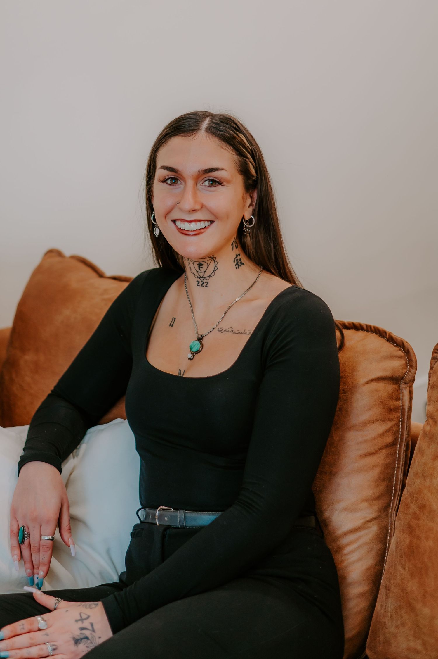 Person in black long sleeve top seated on brown leather couch showing jewelry and tattoo details.