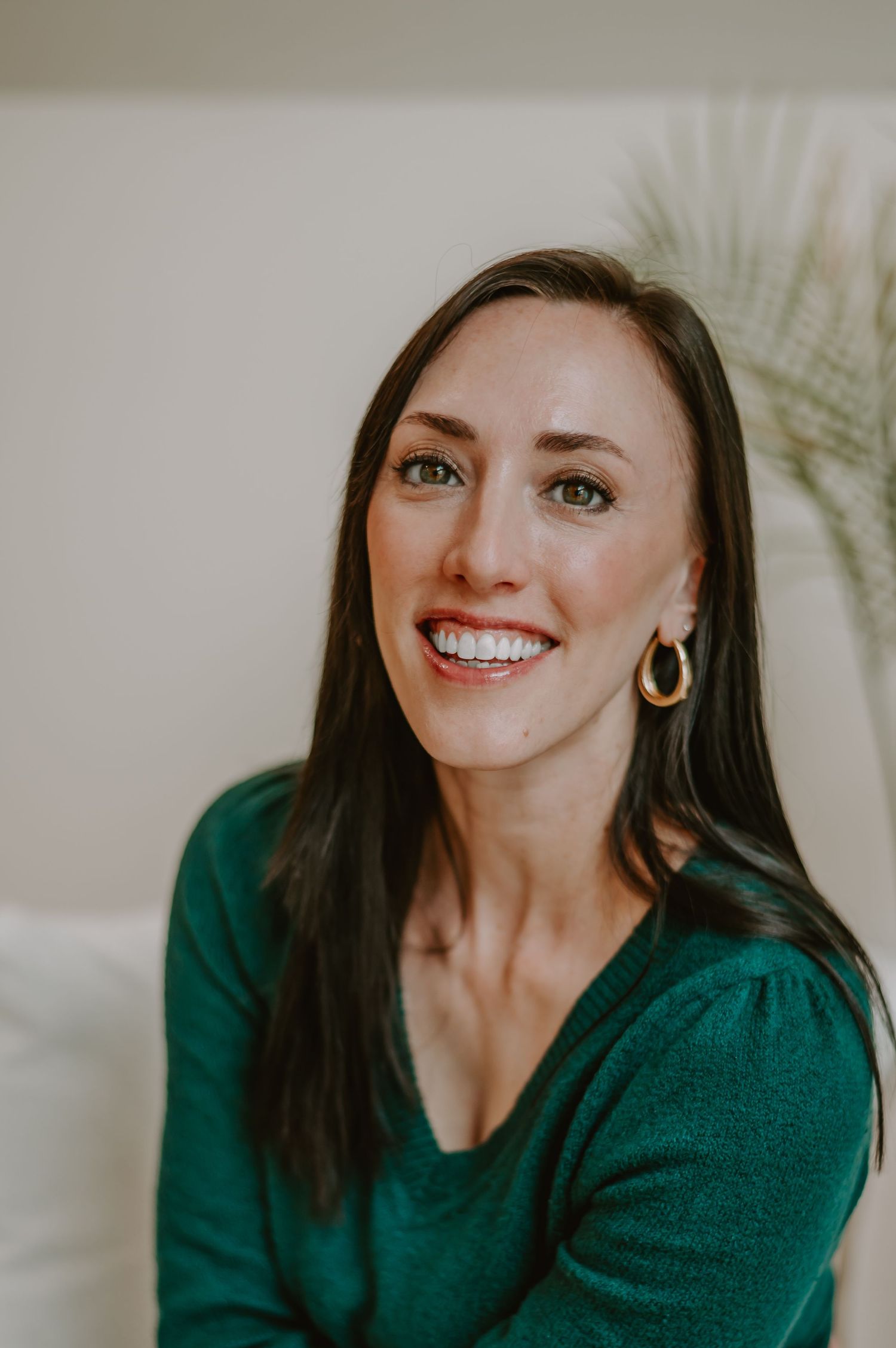 Close-up portraits of someone in emerald sweater smiling against neutral background with palm leaves.