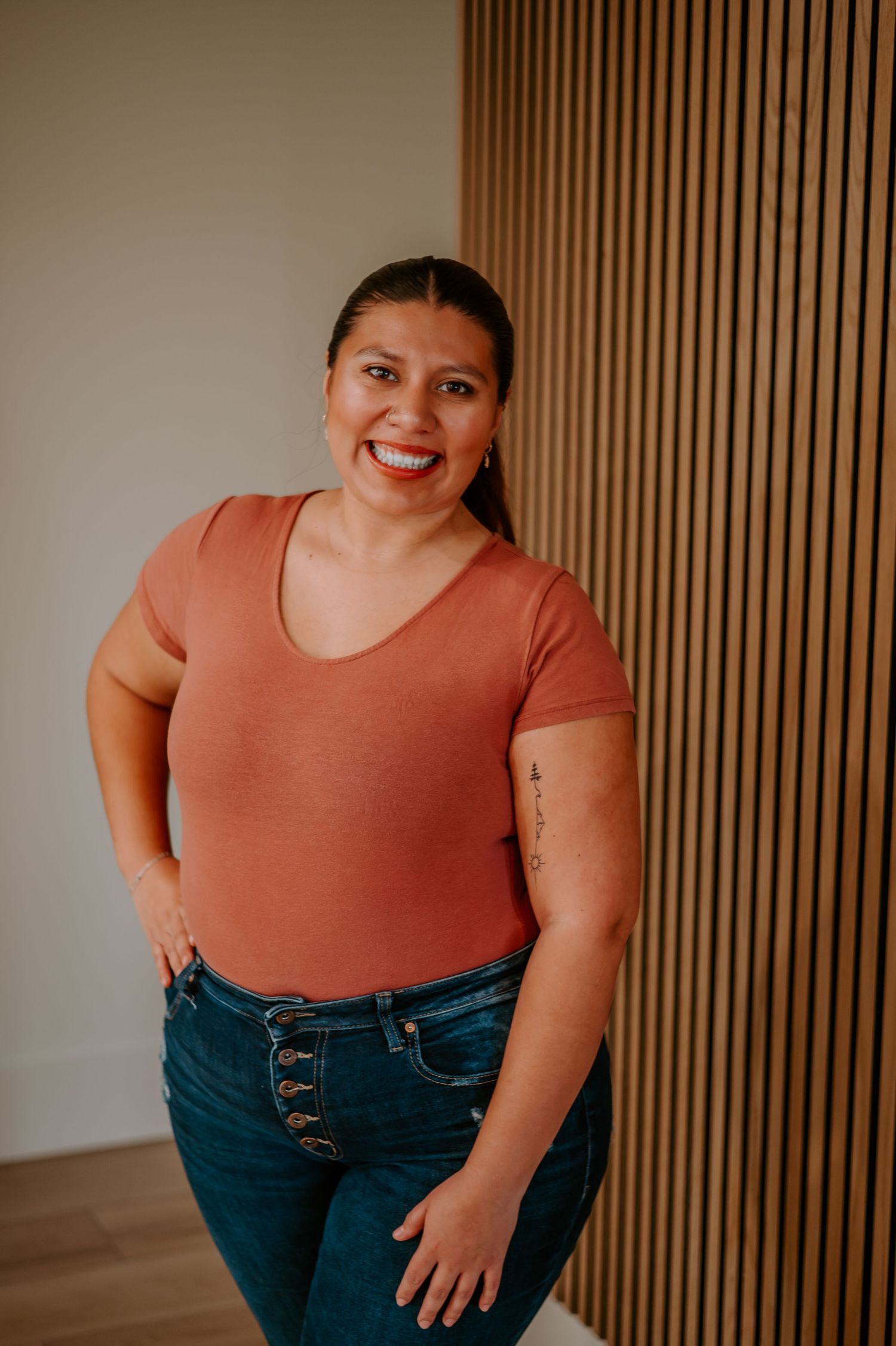 Indoor portrait against wooden slat wall wearing a coral top and button-fly jeans.
