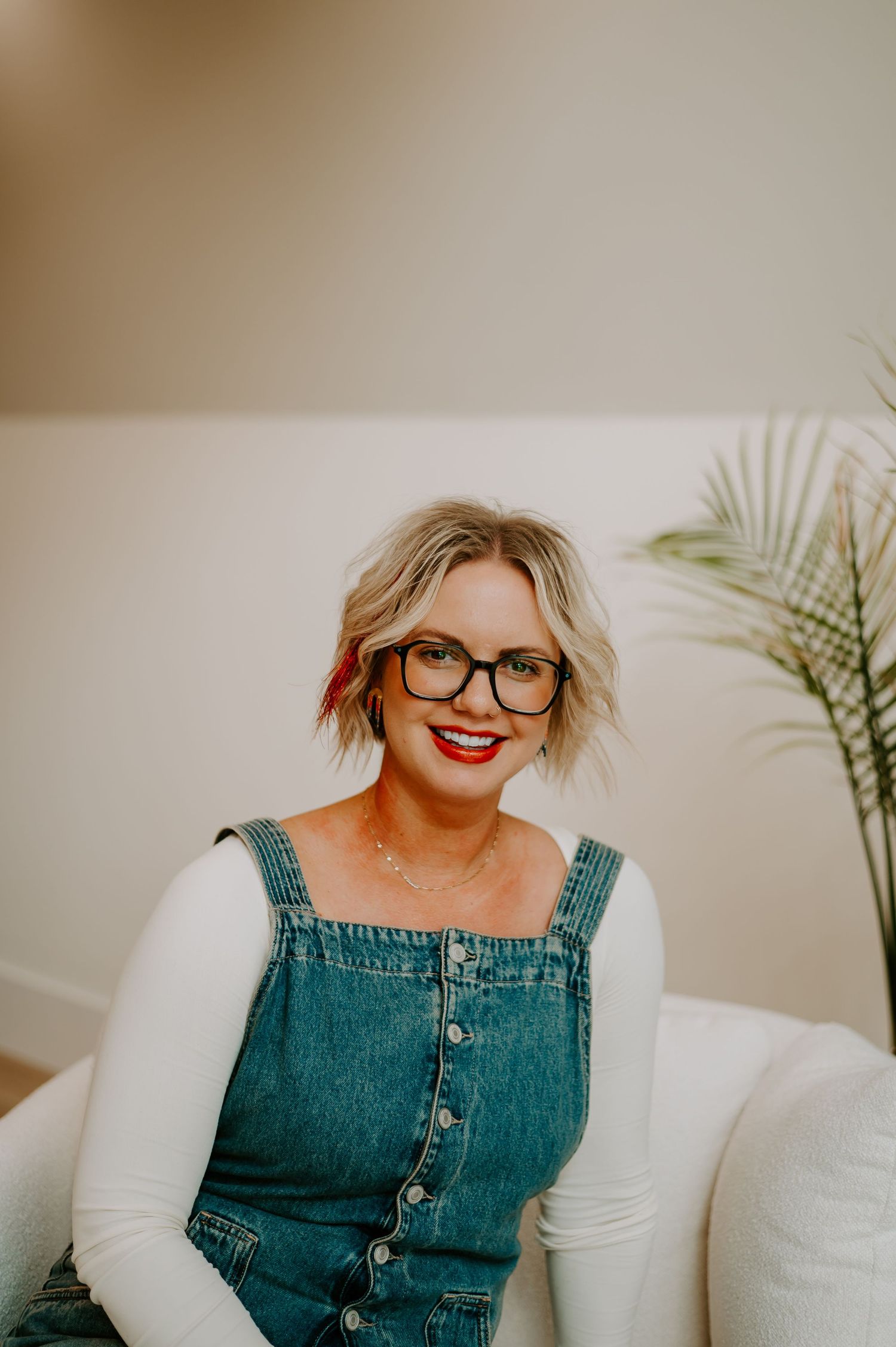 Casual indoor portrait showing a bright smile, glasses and denim overalls against a neutral background.