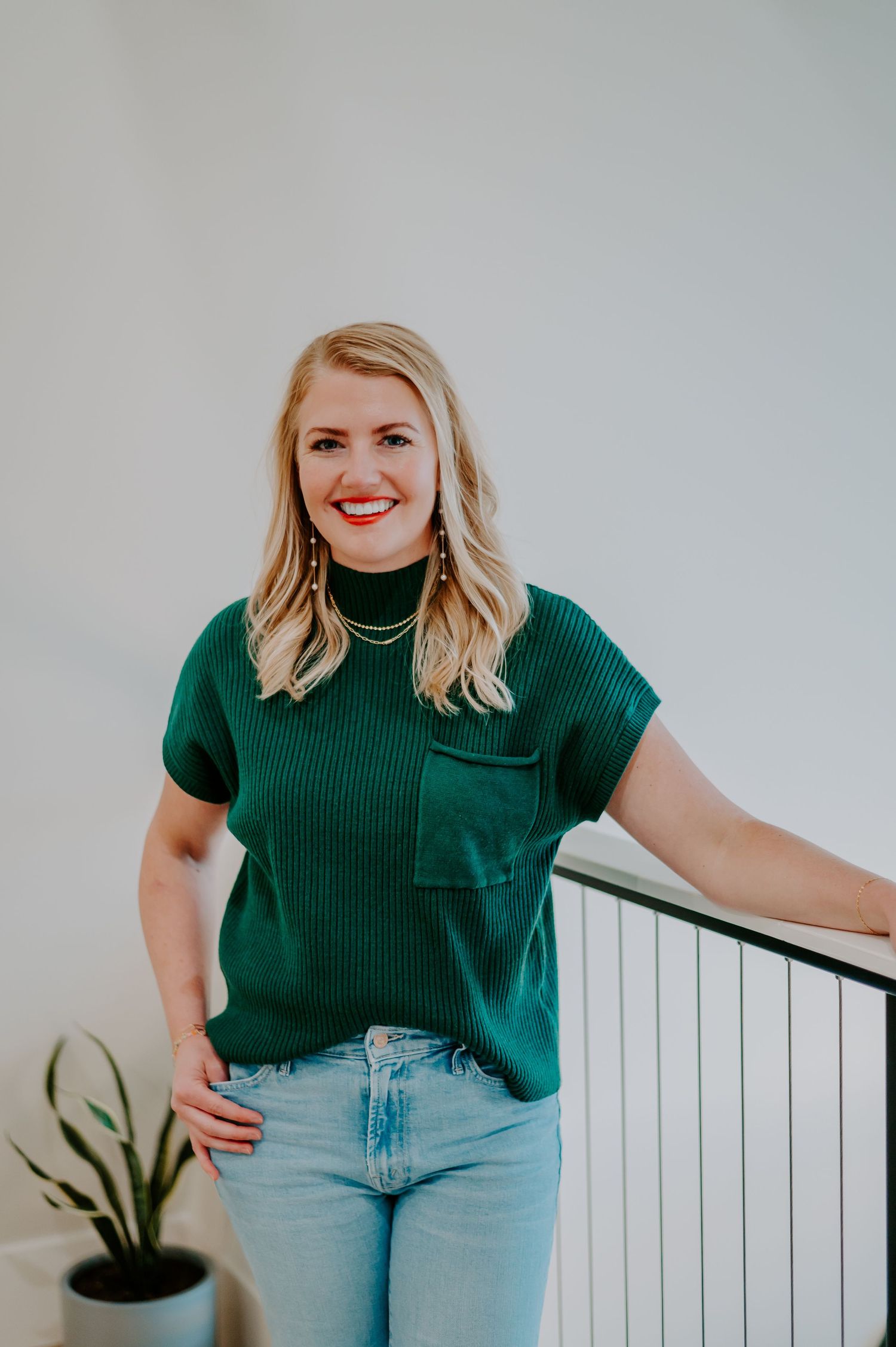 Person in green ribbed top and light jeans posing by white railing.