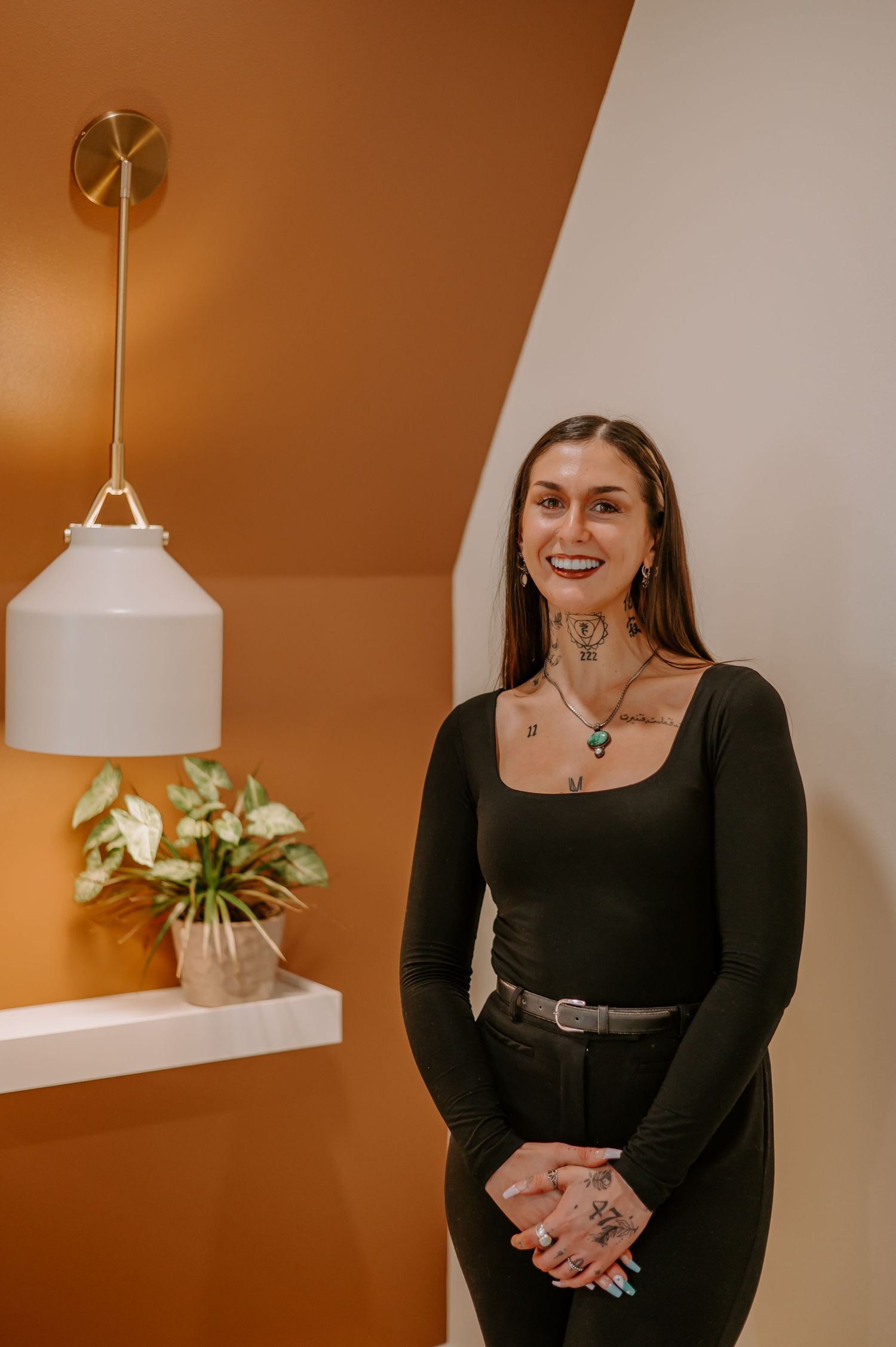 Person in black top posing against orange wall with pendant light and plants.