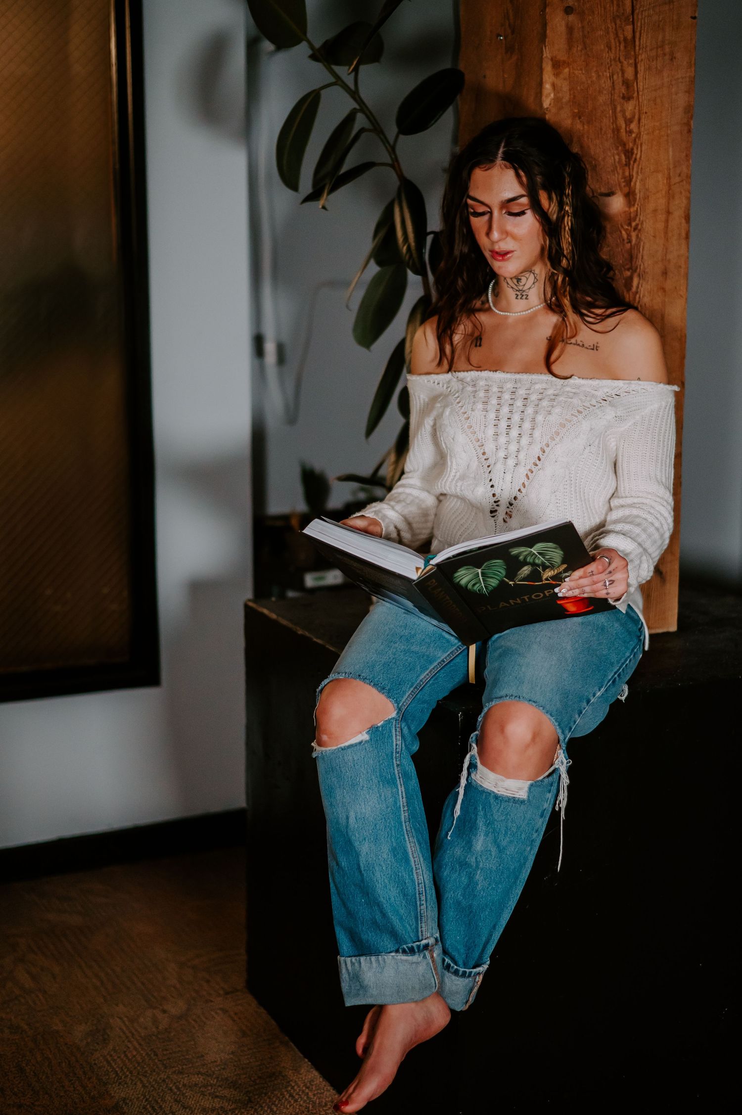 Person in distressed jeans and cream sweater sits casually against wooden wall with potted plant nearby in moody lighting.