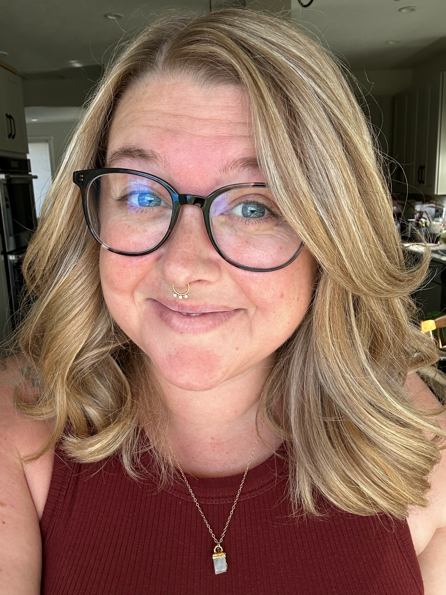 A person with blonde wavy hair and glasses wearing a burgundy top and silver necklace smiles at the camera in a selfie