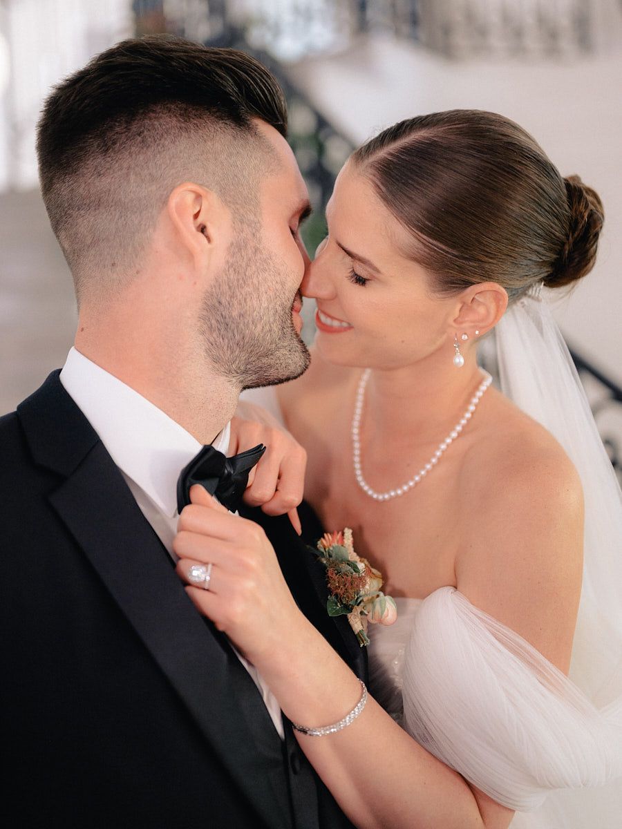 Bride kisses groom while adjusting his bowtie, romantic Riga wedding moment in natural light