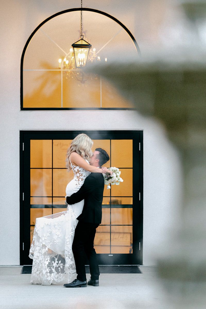 The bride and groom embrace in the soft evening light, with the elegant venue in Latvia behind them.