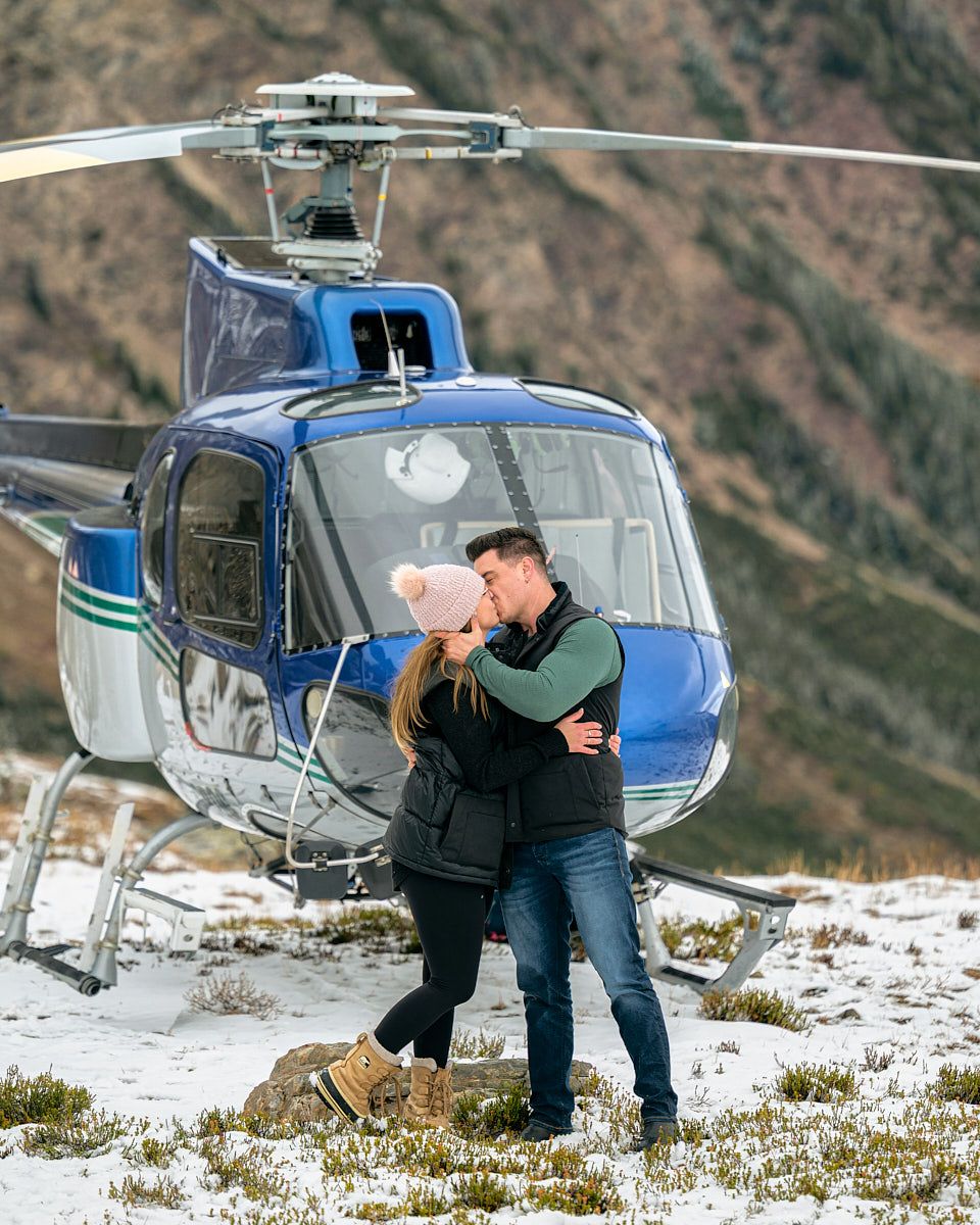 Engaged couple sharing a kiss next to helicopter, adventurous engagement session in Europe
