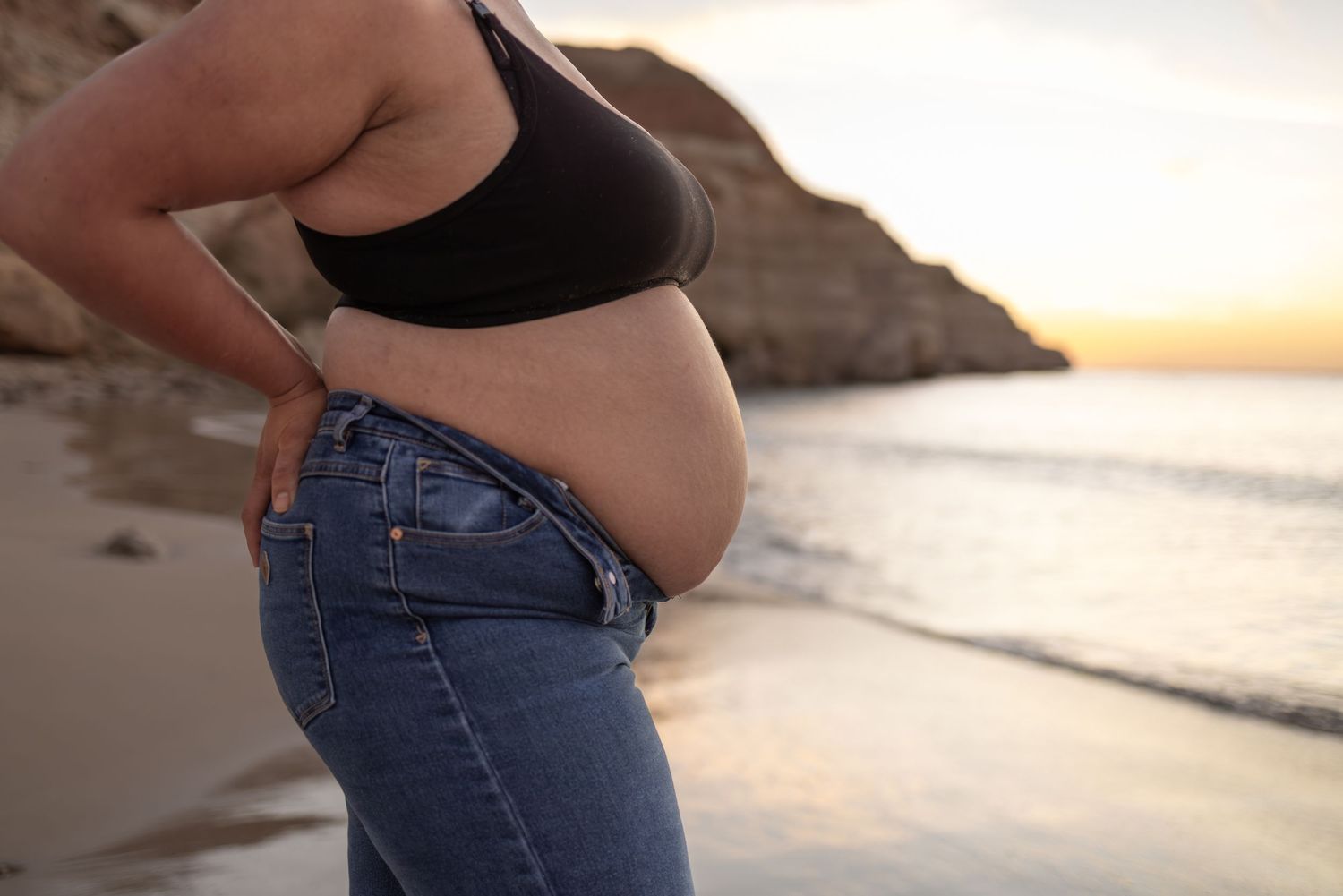 Side view of pregnant belly in black bra and jeans against sunset beach backdrop.