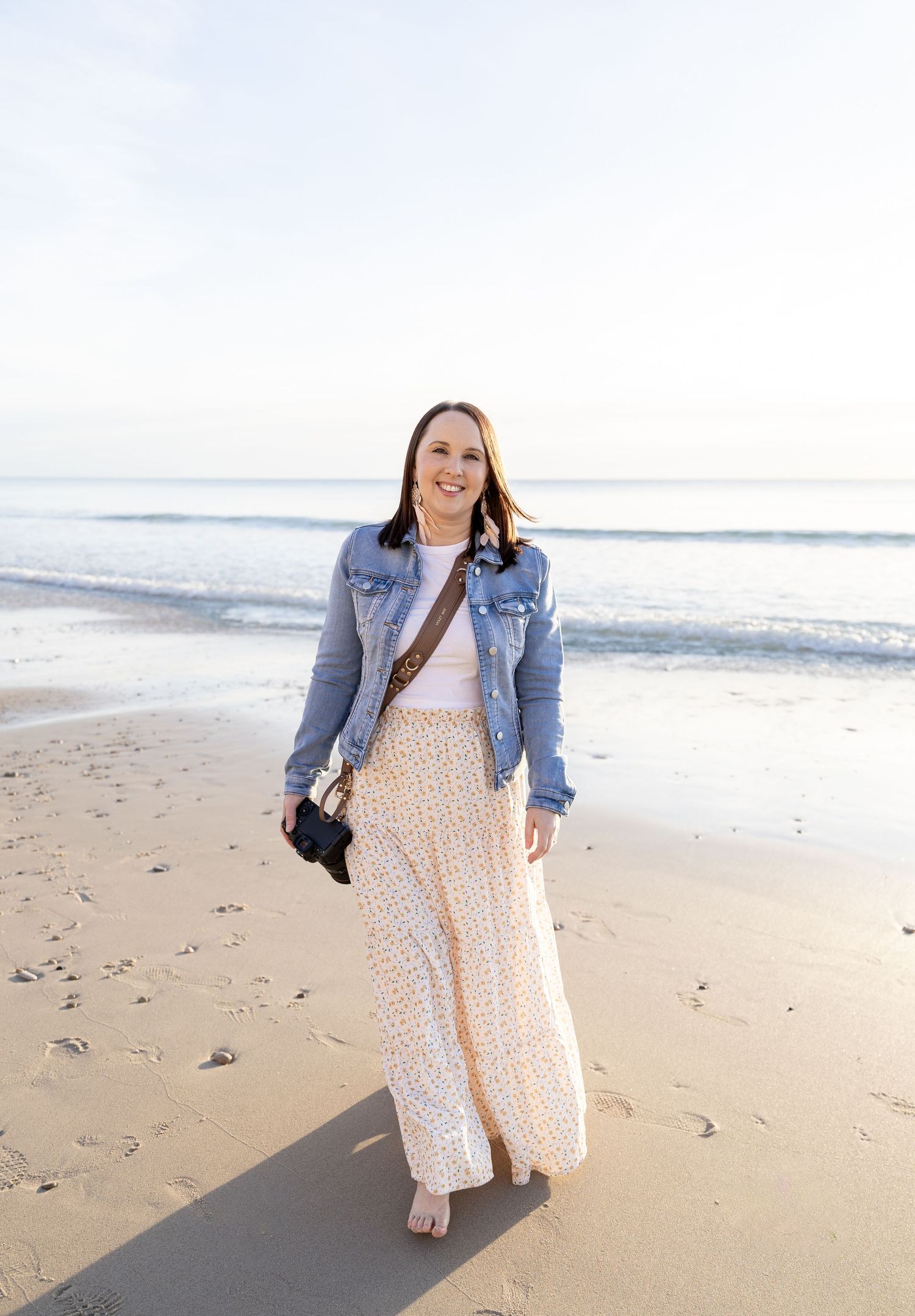 Adelaide Photographer Kelly-Jade Schinella walks barefoot on beach wearing denim jacket and flowing floral maxi dress.