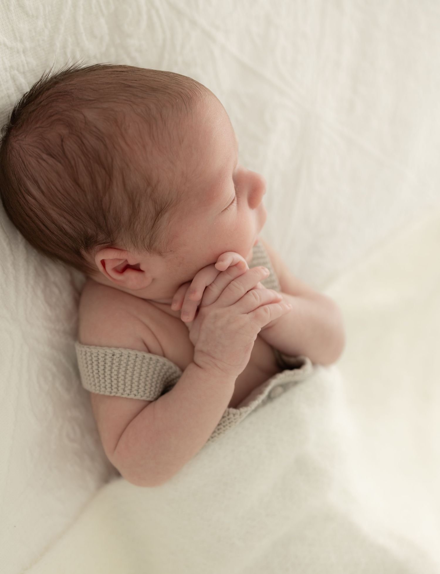 Newborn baby in neutral tones curled up sleeping on white textured backdrop.