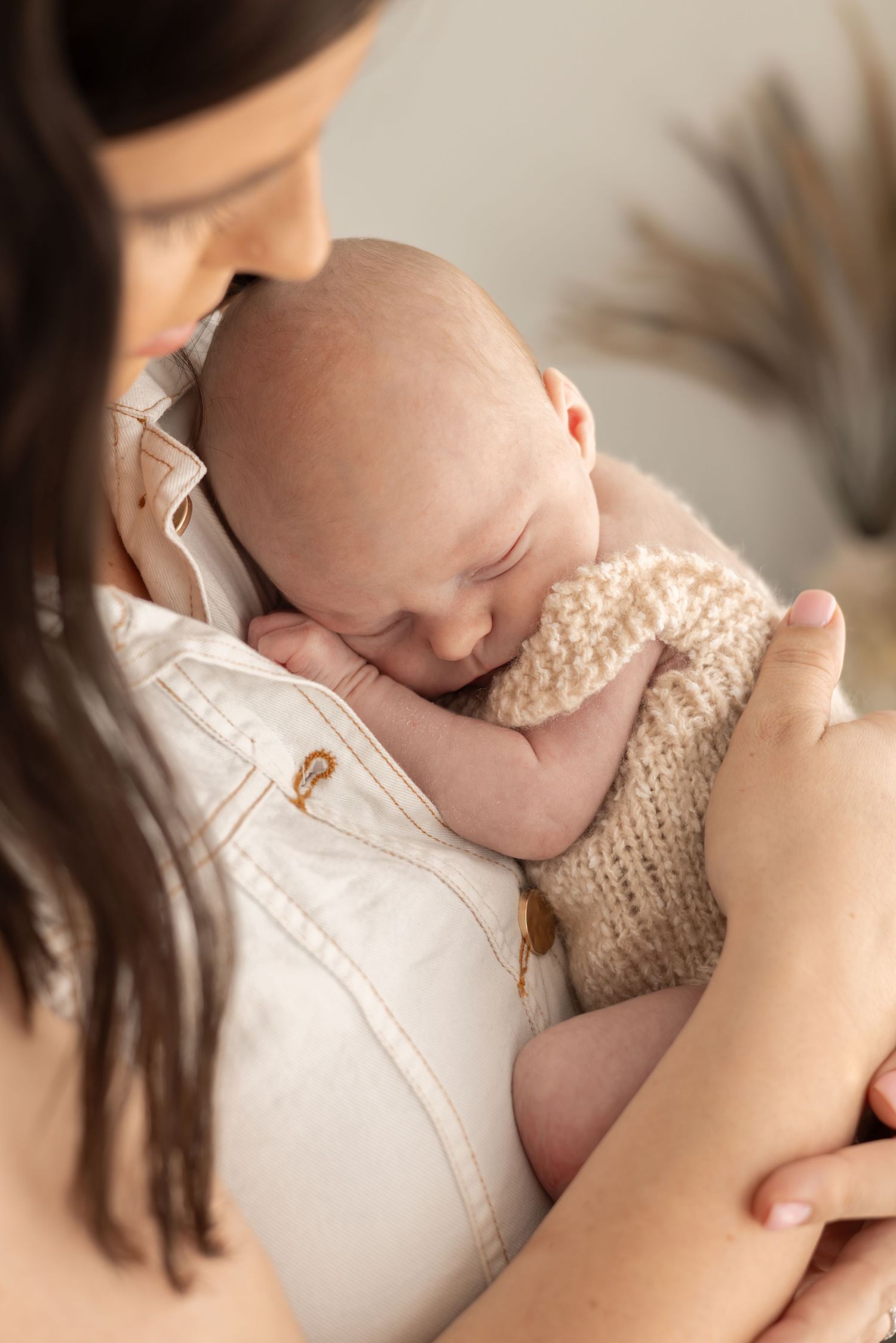 Close-up of sleeping newborn with cream knit romper being cradled against mothers chest with white clothing.