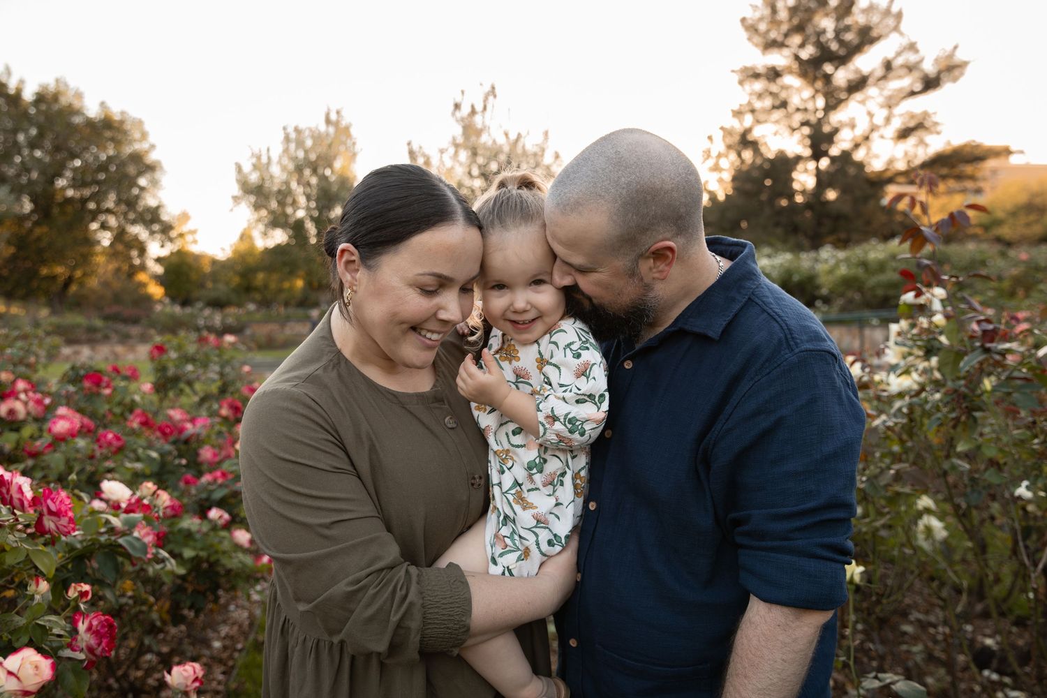 Family of three sharing a tender moment in a rose garden at golden hour, parents holding toddler close.