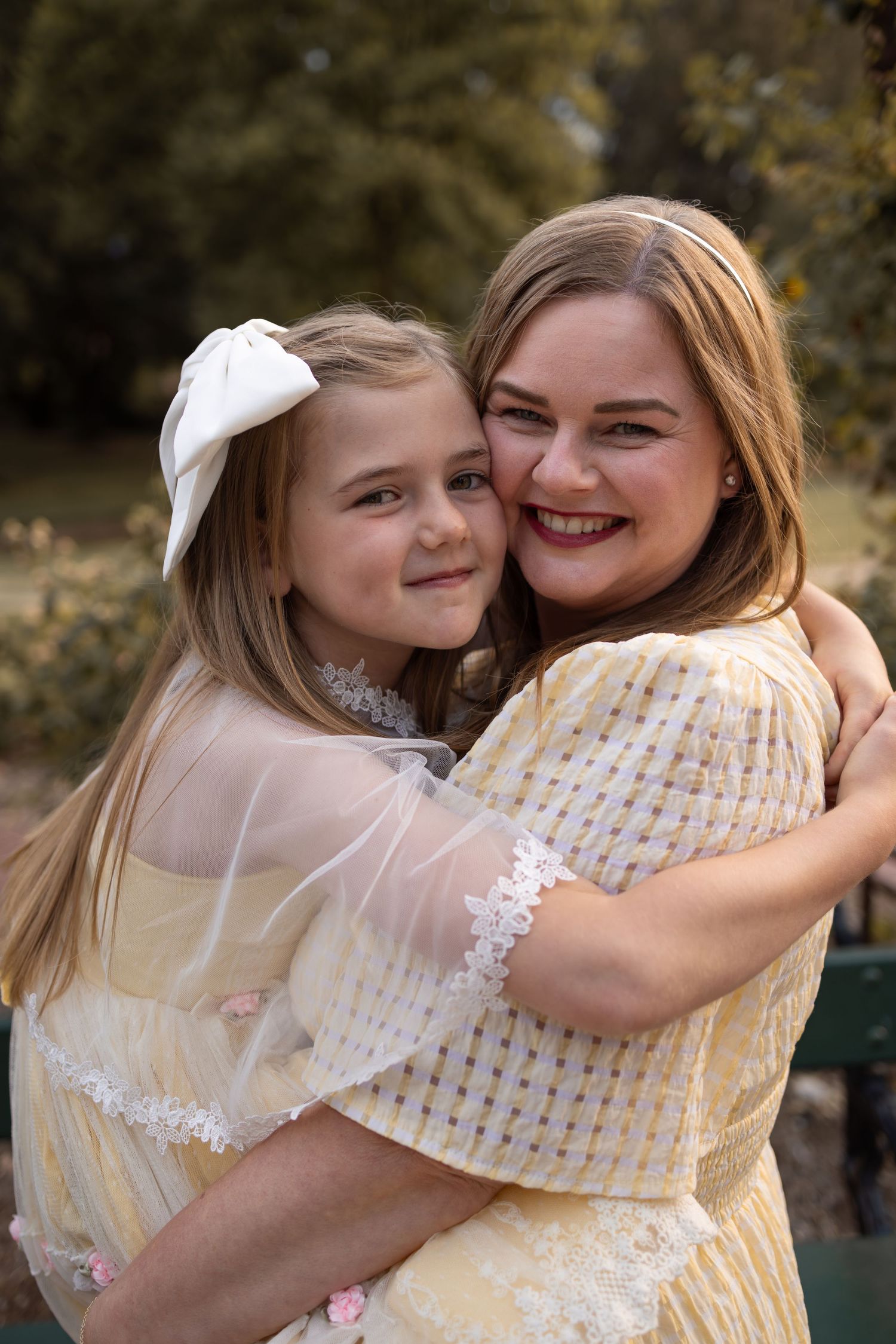 A smiling woman and young girl in matching yellow dresses share a warm hug outdoors, with the girl wearing a white bow.