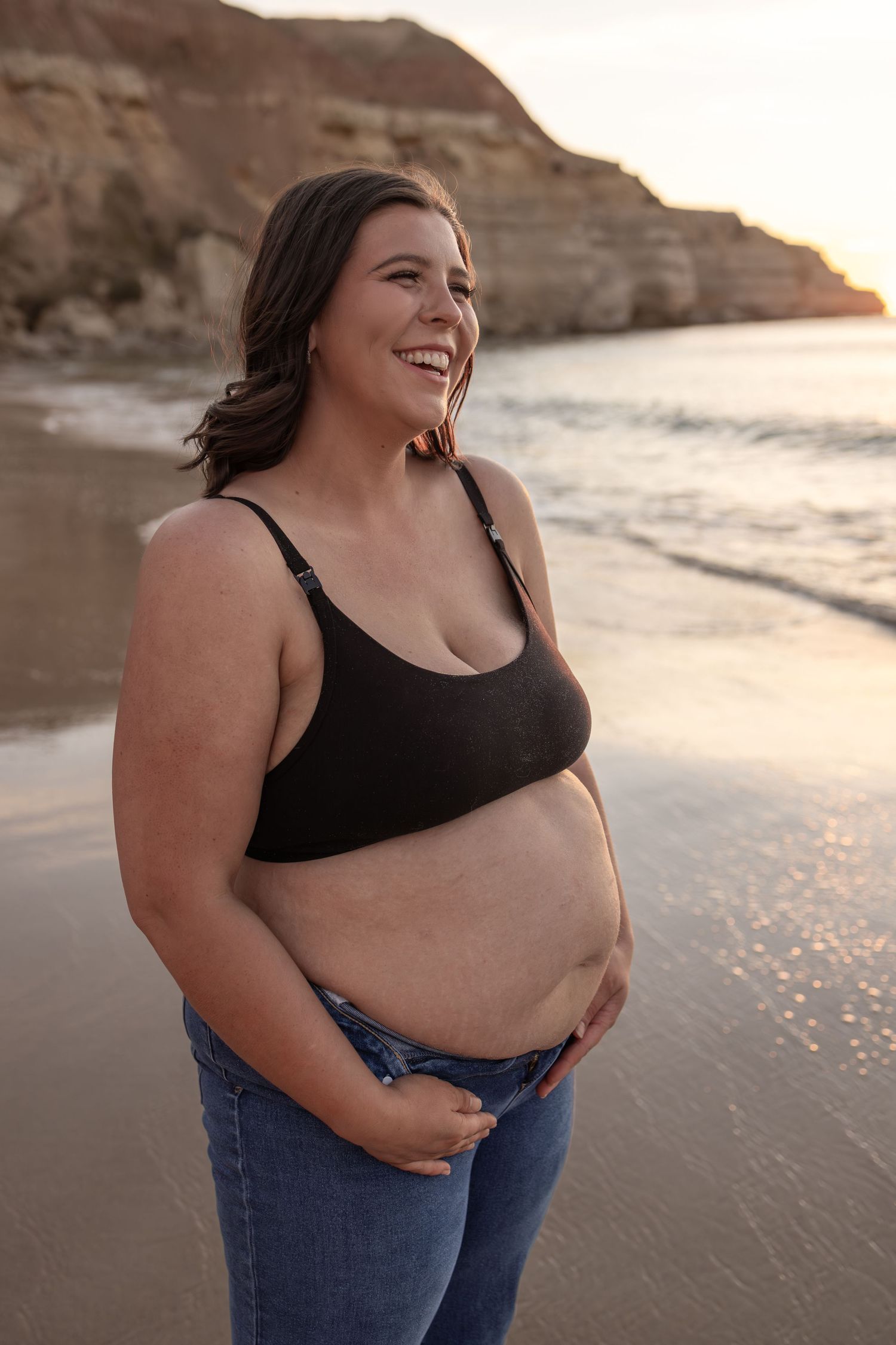 Joyful expectant mother in black bra and jeans poses on beach at golden hour.