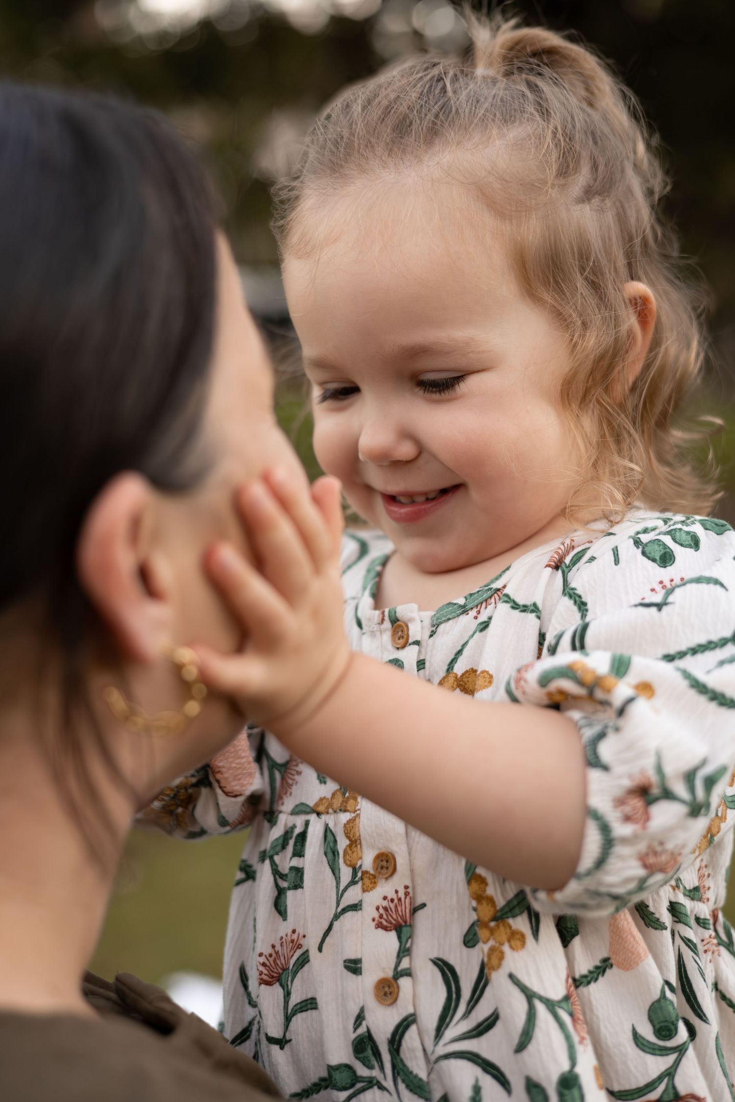 A smiling toddler girl in a floral dress gently touches a woman's face outdoors.