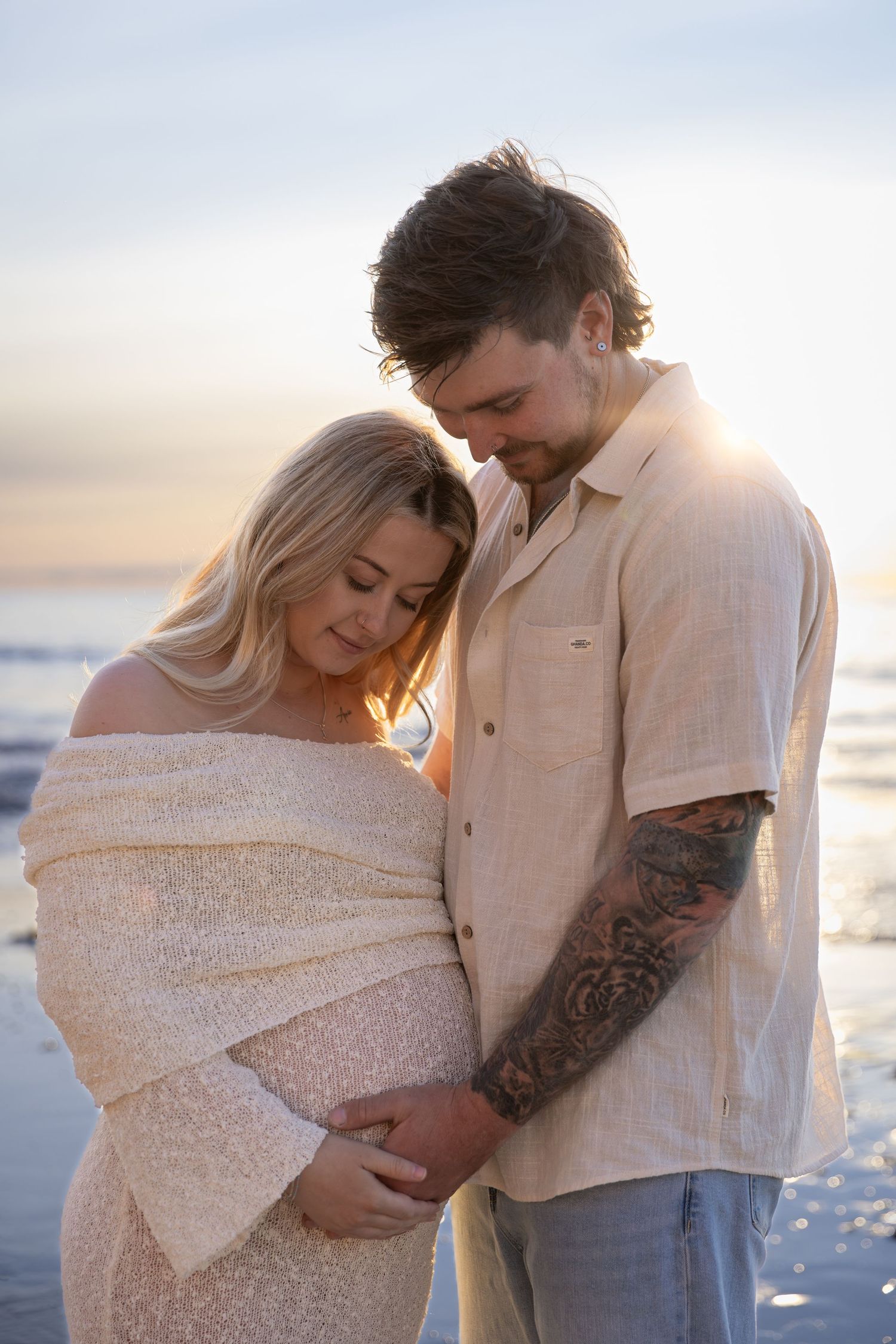 Romantic beach maternity photos of couple embracing at sunset in neutral tones.