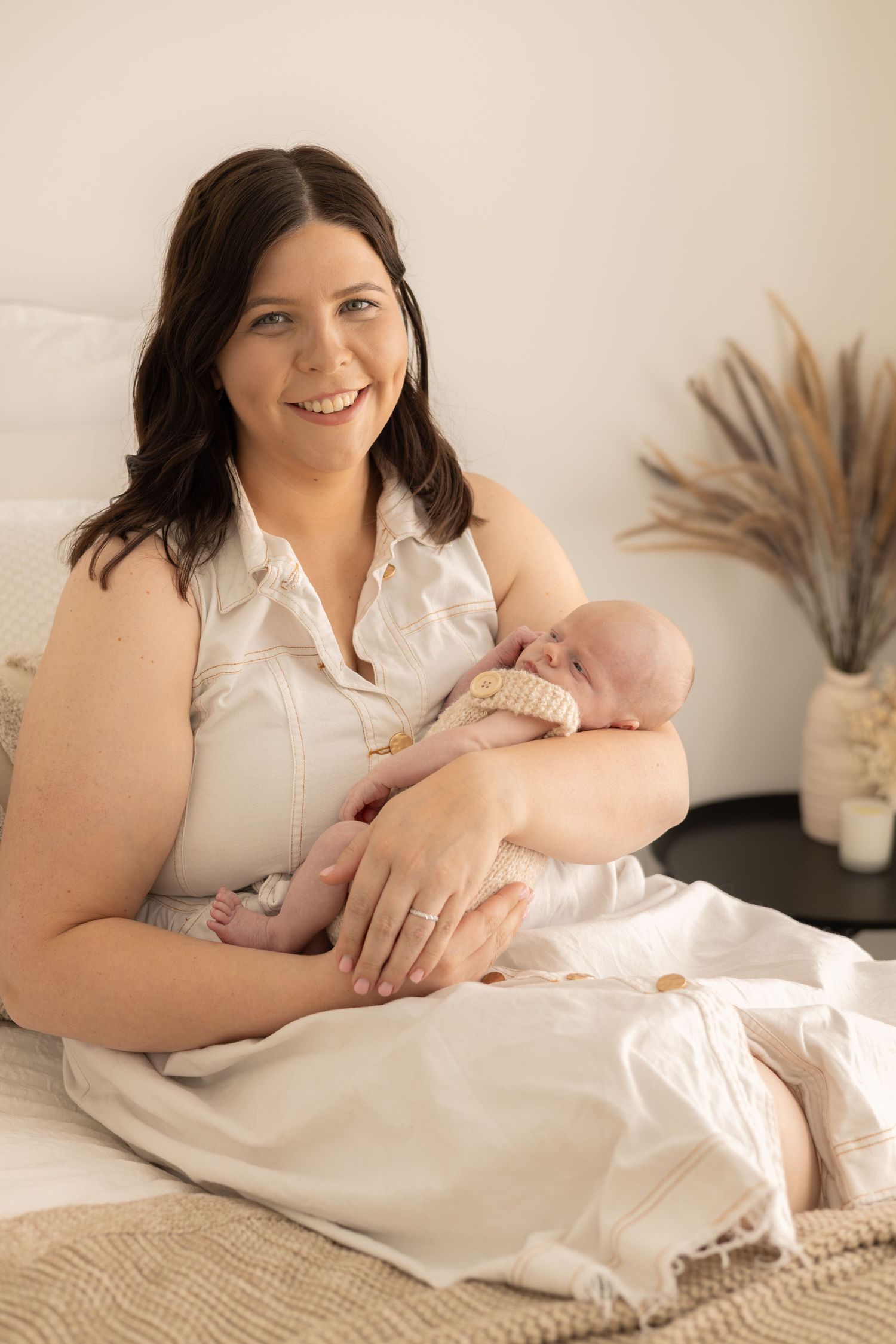 Mother in white dress holds sleeping newborn on cozy bed with neutral decor.