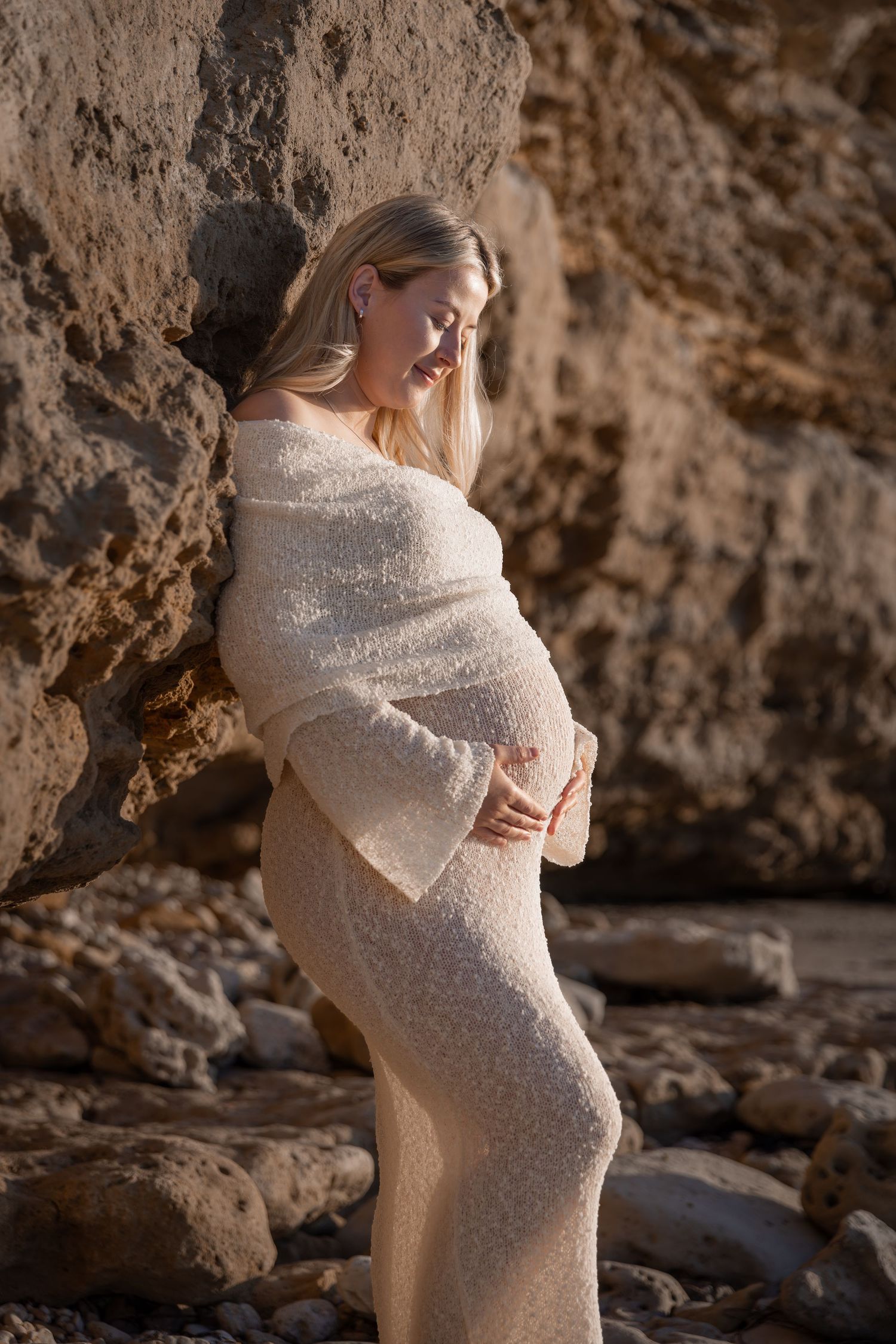 Elegant maternity portraits against rocky backdrop in stretchy knitted white dress.