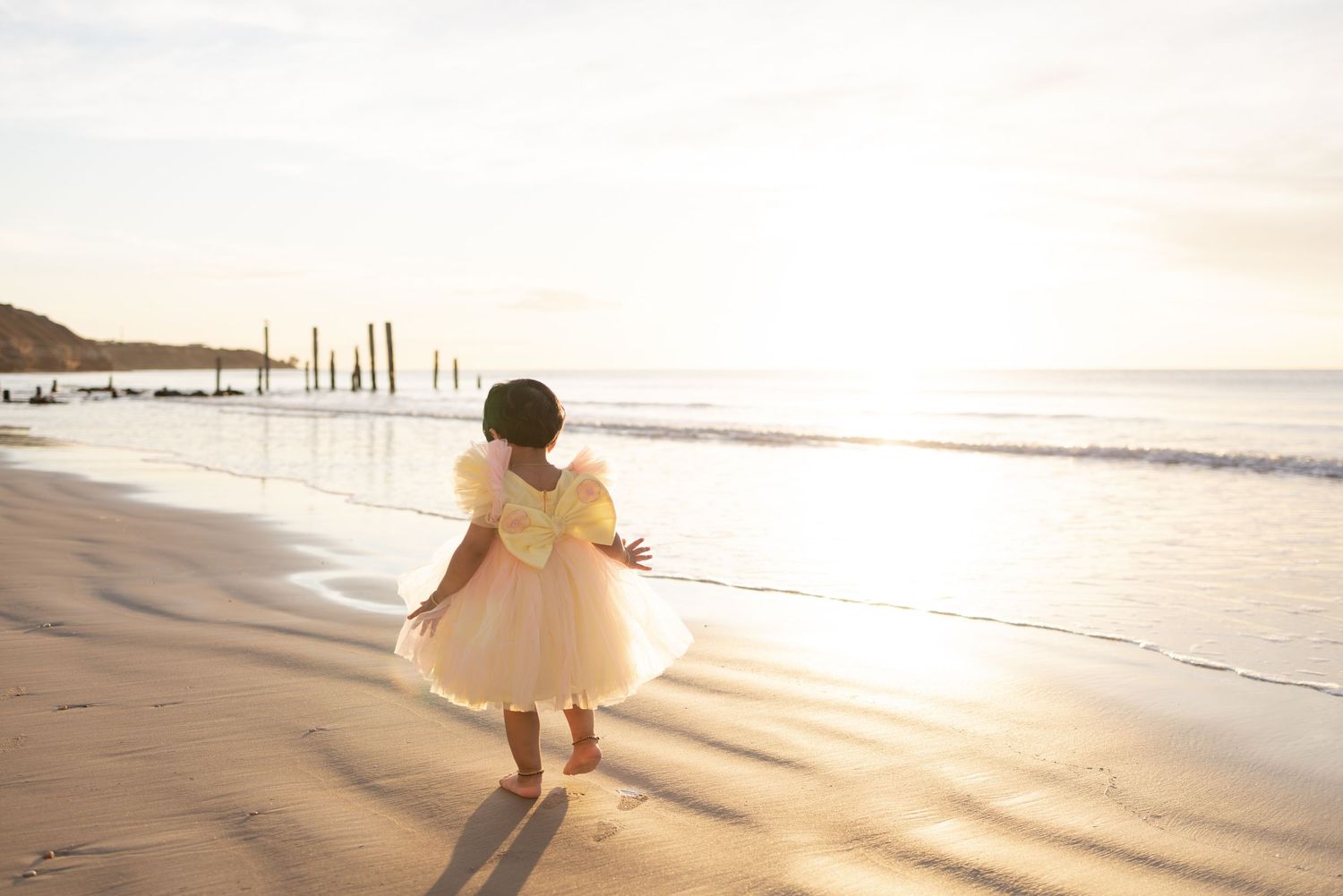 Little girl in pink and yellow dress walks along Port Willunga beach at sunset with ocean waves in background.