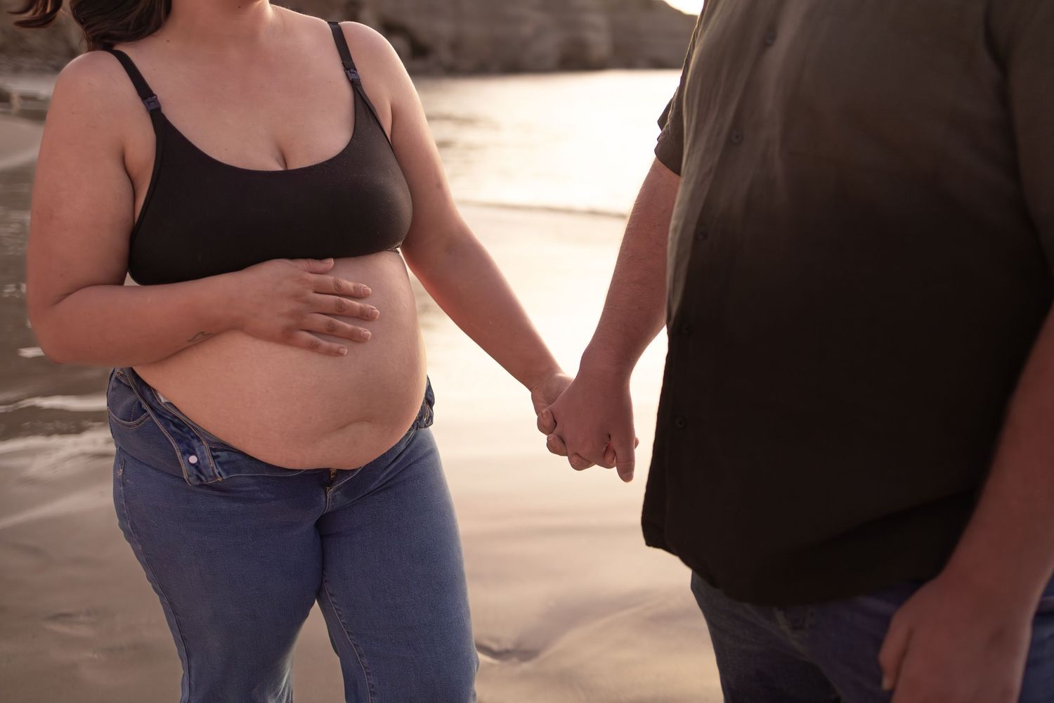 Couple holds hands during beach maternity photoshoot at sunset.