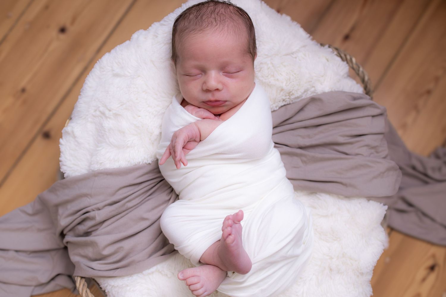 Newborn baby swaddled in white fabric resting peacefully in wicker basket on wooden floor.