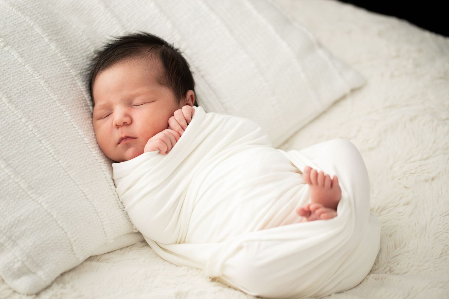 Sleeping newborn wrapped in soft white blanket against textured white background.