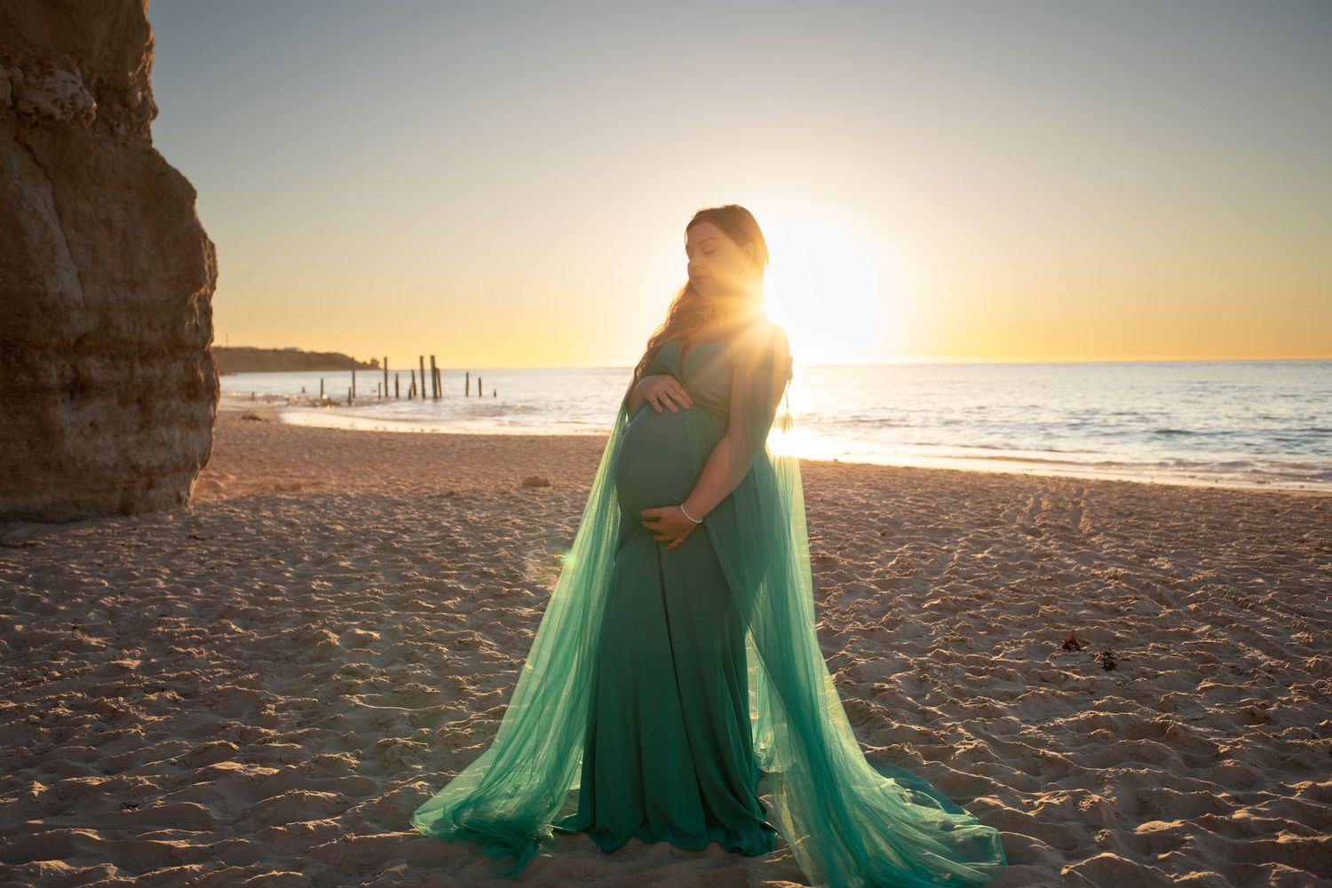 A pregnant mother draped in a flowing green chiffon dress which catches the light during a sunset maternity photoshoot on a sandy beach.