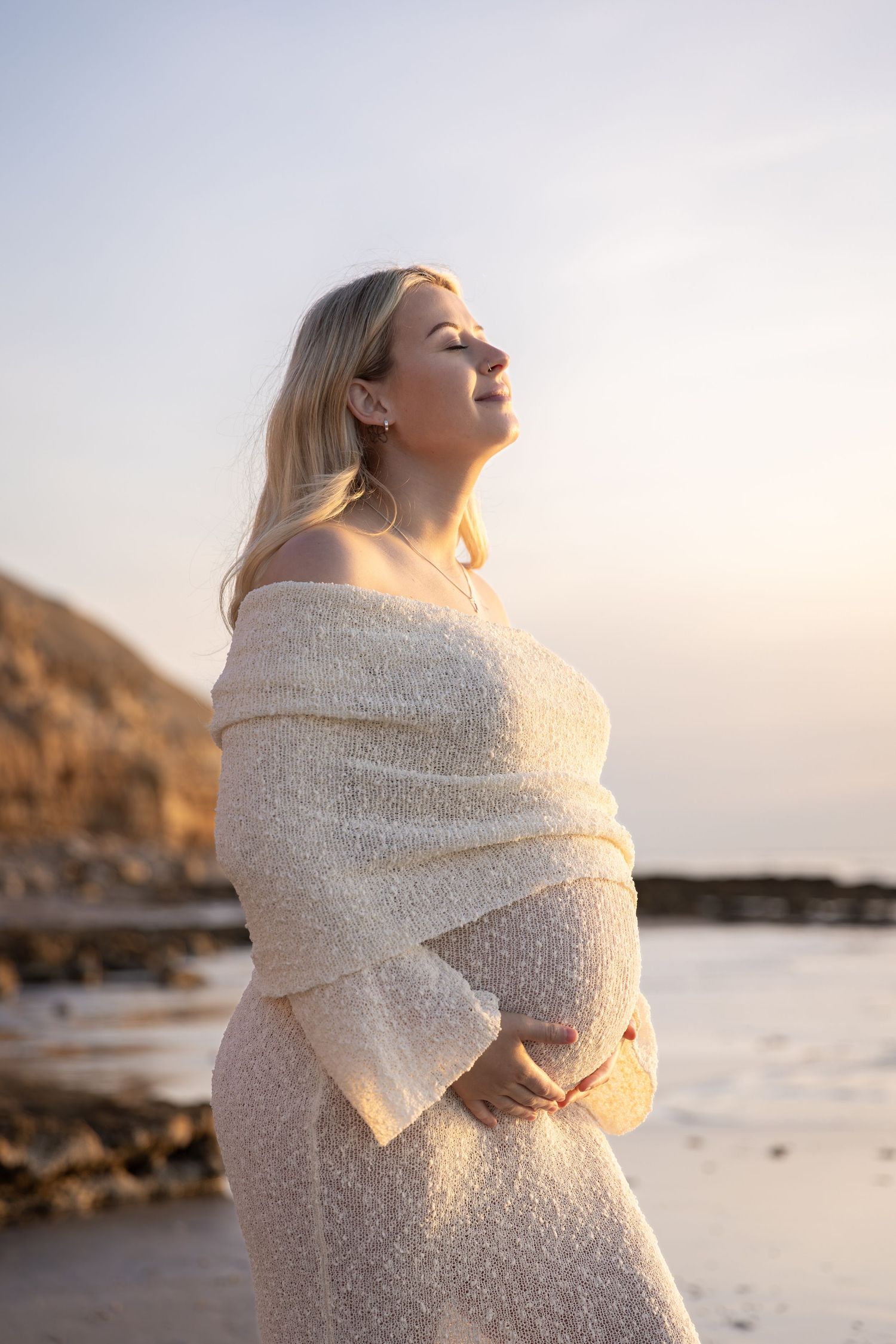 Pregnant woman wrapped in white knit shawl poses on beach at sunset.