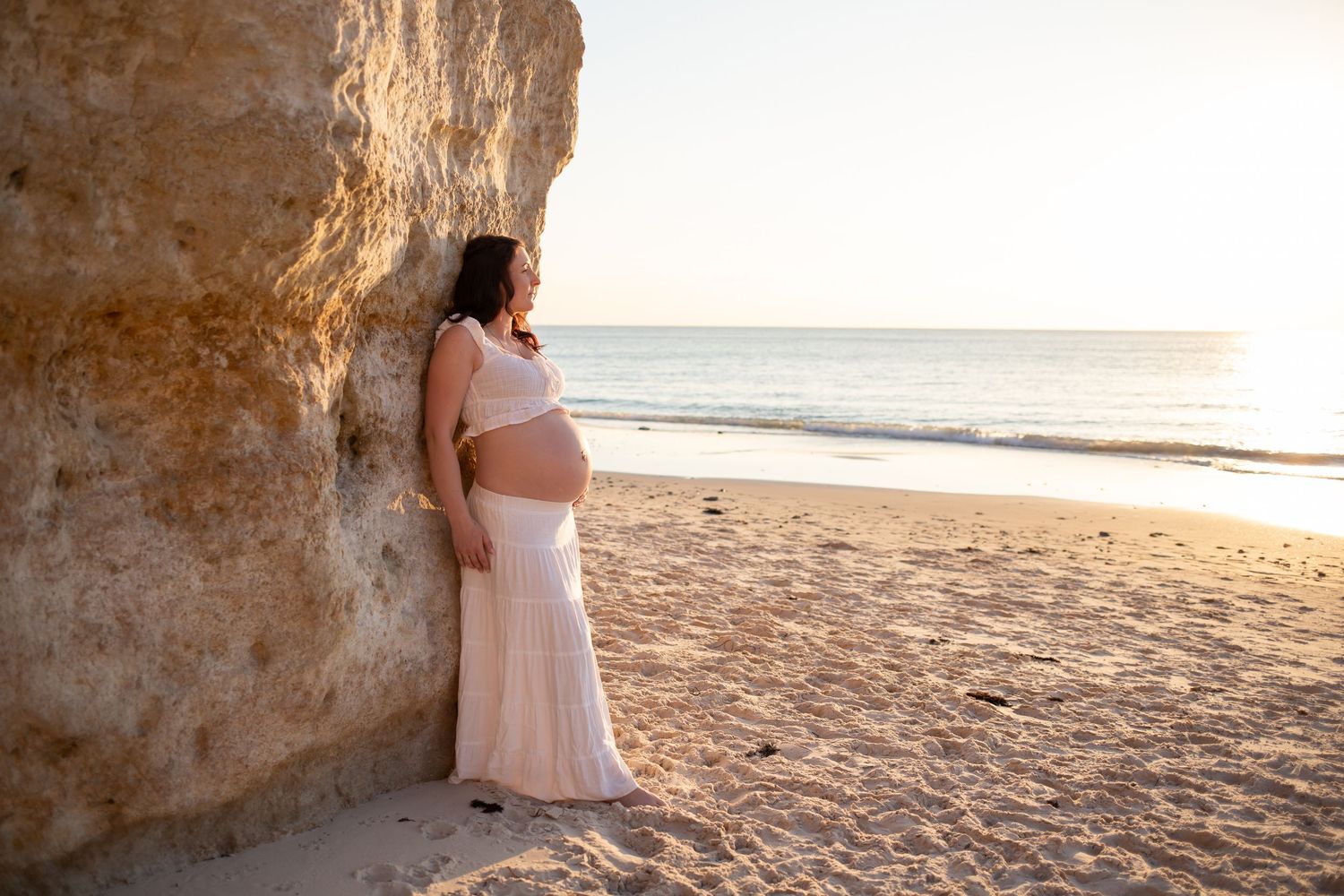 Maternity photo of mother in white skirt and top against rocky beach backdrop at sunset.