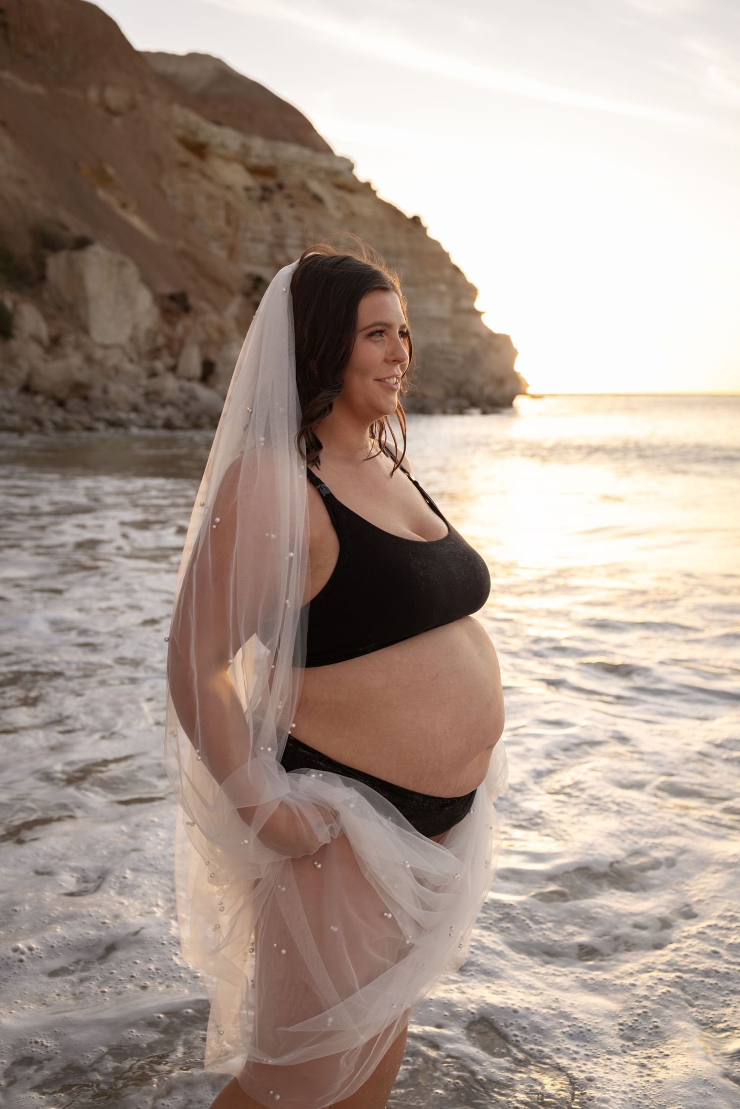 Expectant mother in black bra and veil poses on beach with cliffs and sunset behind.