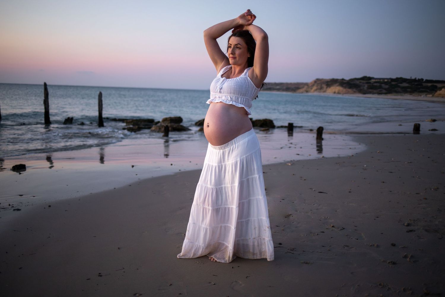 A pregnant woman in a white dress poses gracefully on a beach at blue hour with wooden jetty posts in the background.