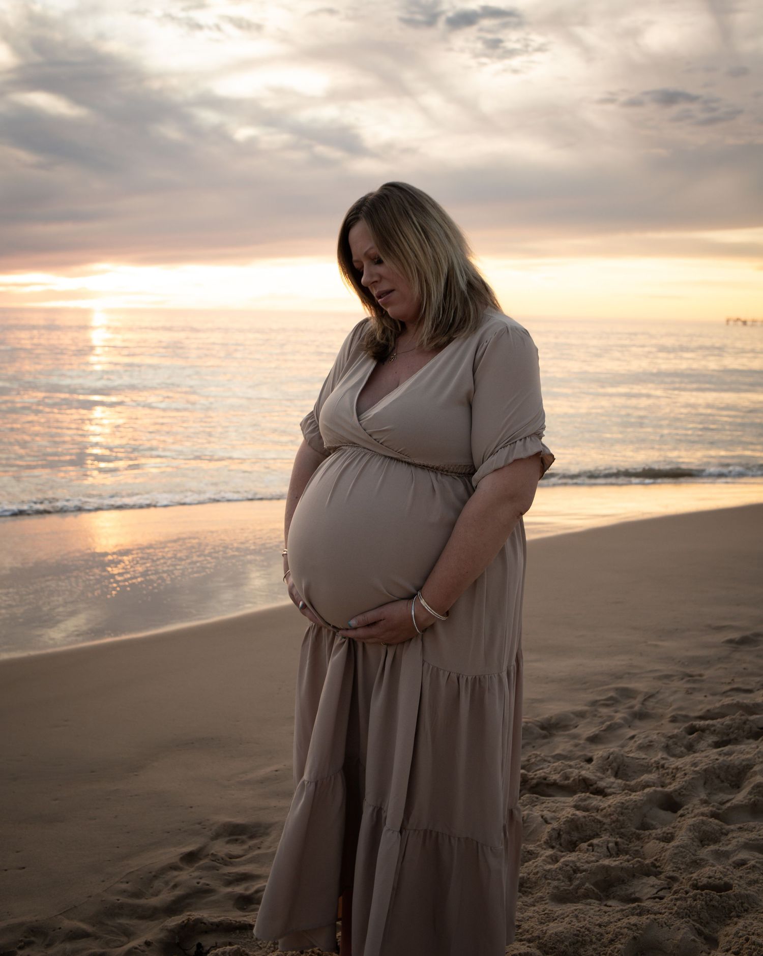 Expectant mother in flowing dress stands on beach during golden hour sunset.