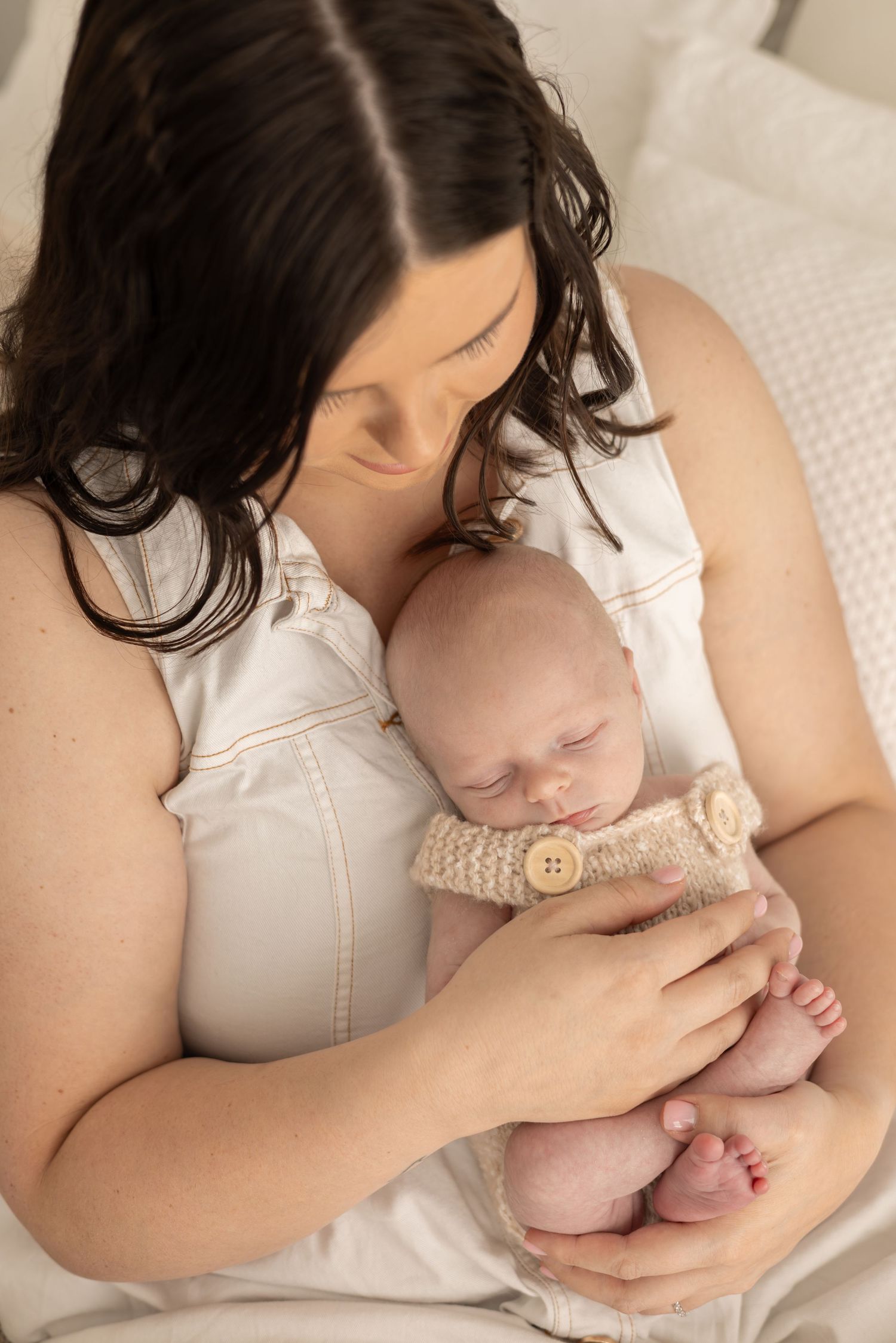 Newborn baby wearing knit romper being held gently against mothers chest on white bedding.