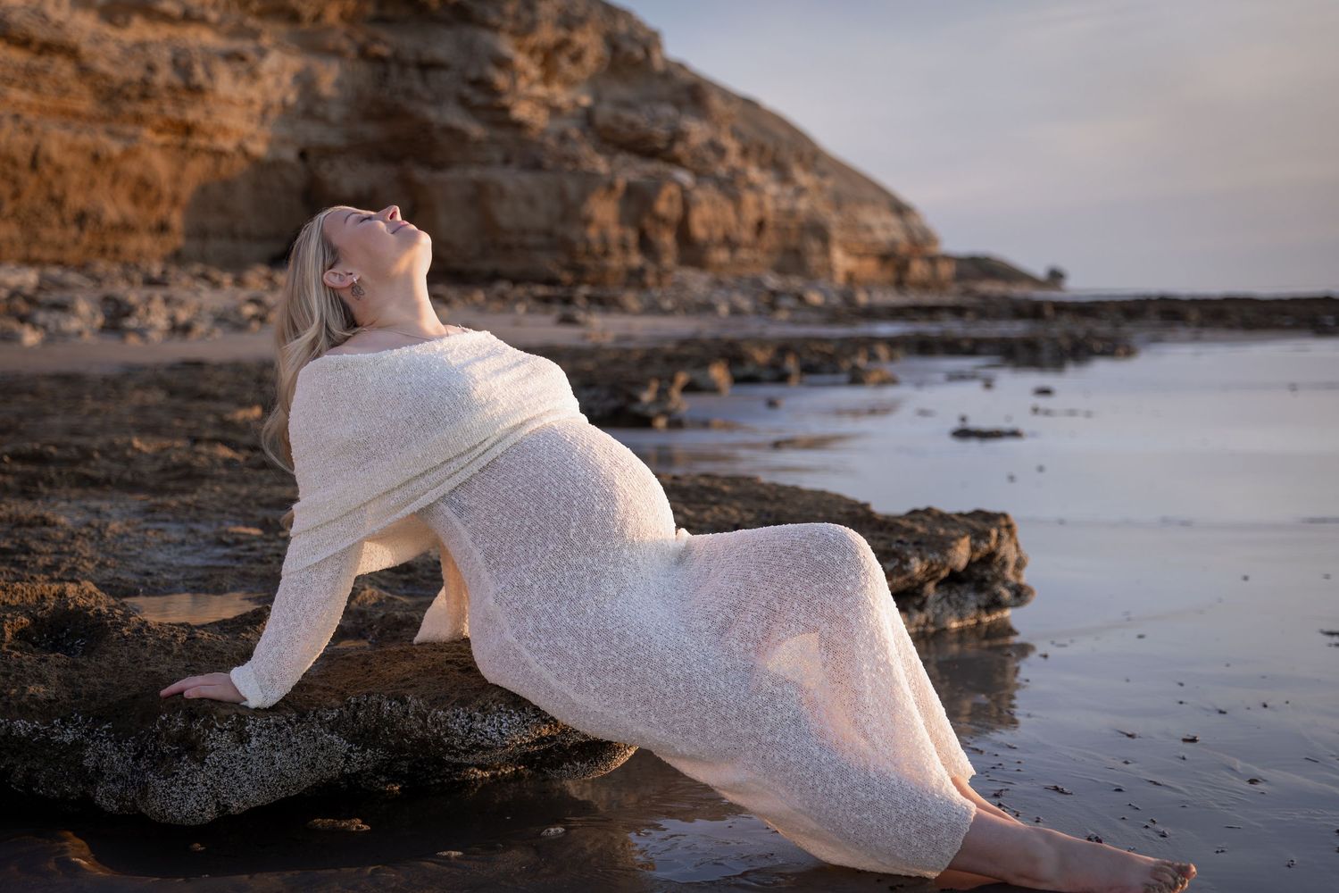 Maternity photos on rocky beach with pregnant woman in flowing white dress at sunset.