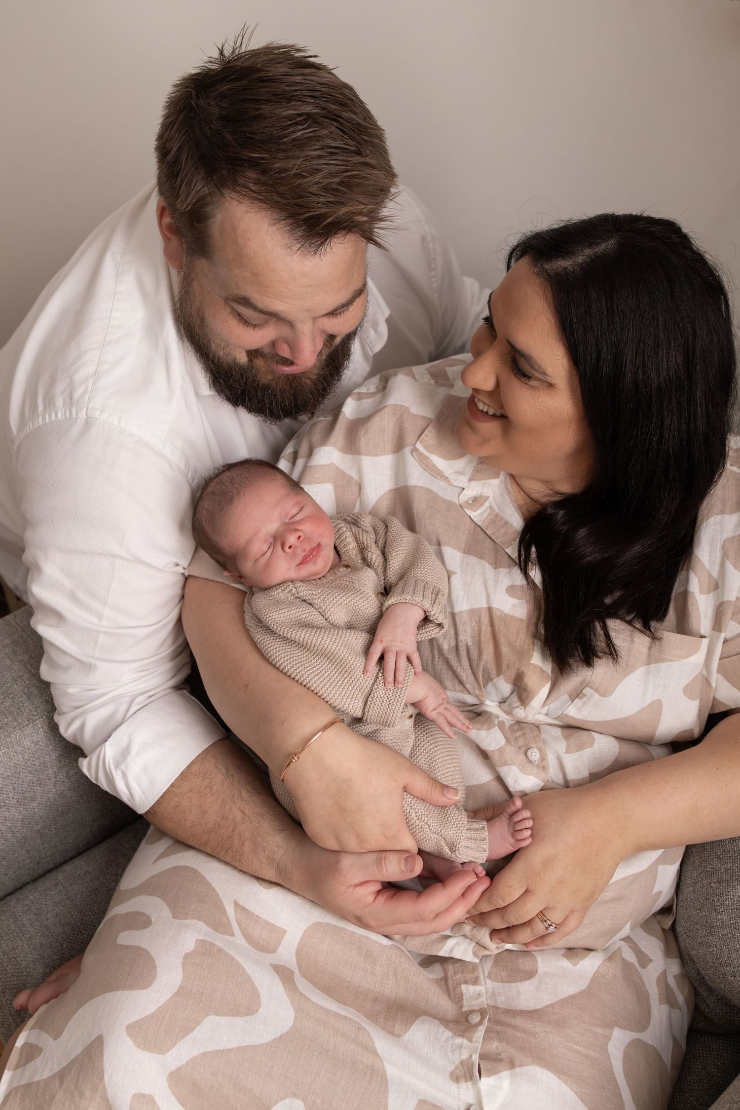A tender family moment as parents cradle their newborn baby on a grey armchair.